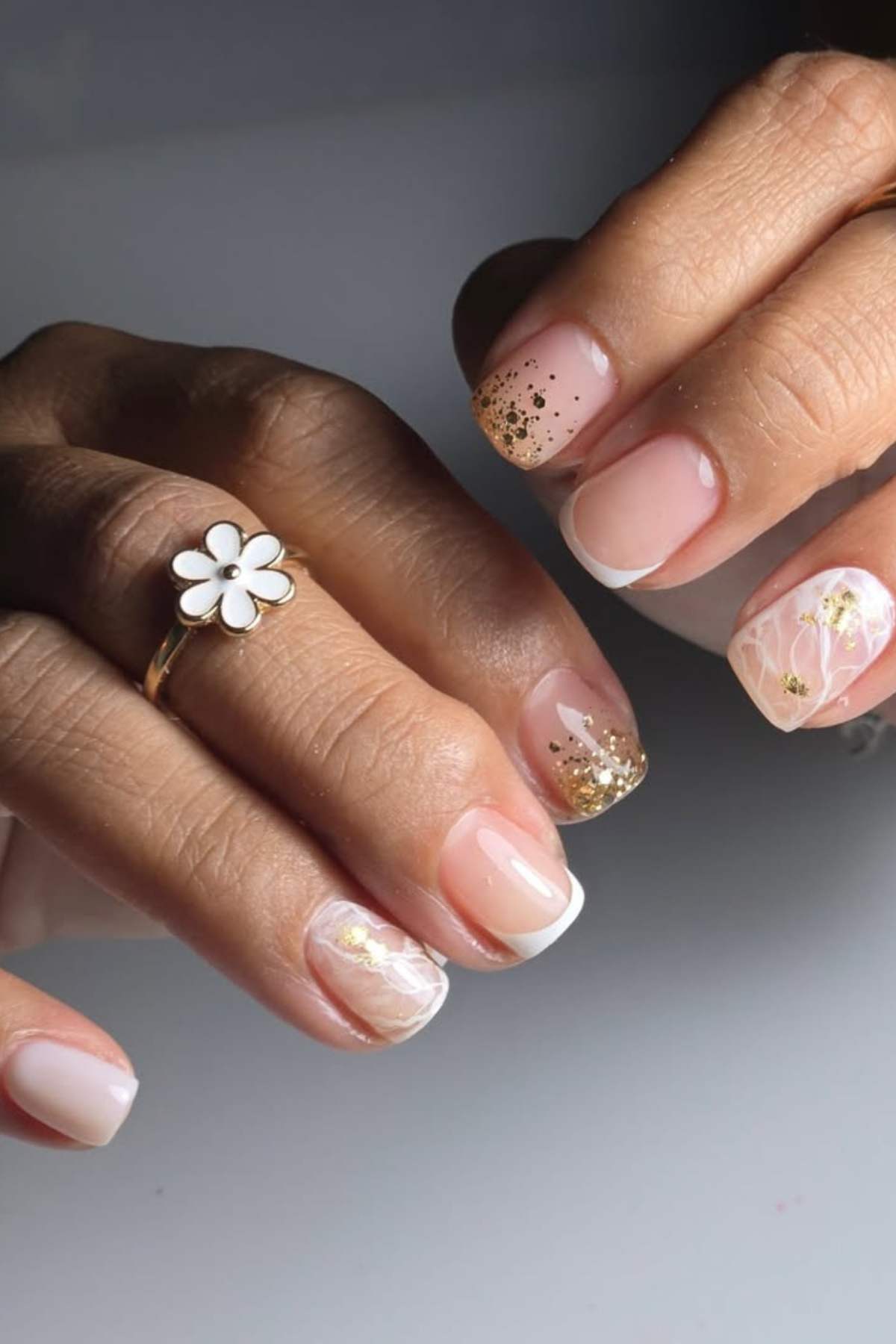 Close-up of hands with a gold flower ring and manicured nails featuring a pink and white French tip, gold glitter, and floral nail art designs.