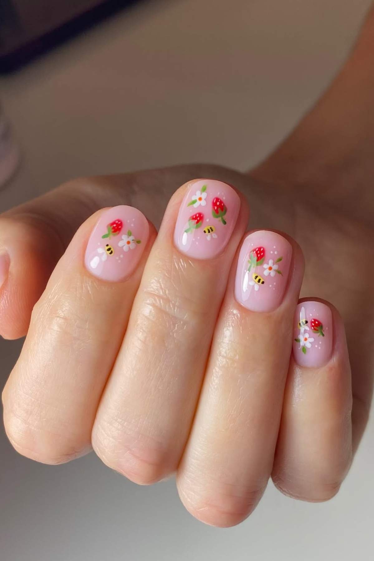 A hand with short nails painted with light pink polish, featuring small, detailed strawberry, bee, and flower designs on each nail.
