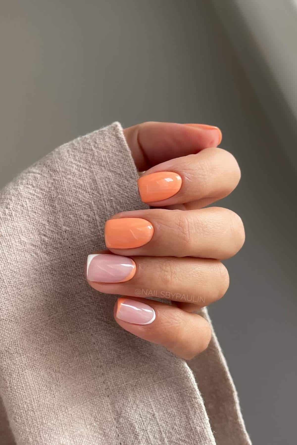 A hand with short, square-shaped nails featuring a glossy peach color on four fingers and a light pink accent nail with a white outline.
