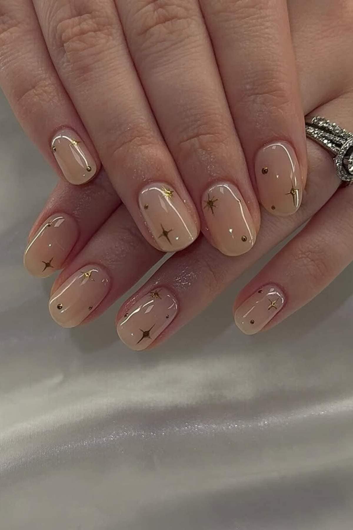 Close-up of hands with short, almond-shaped nude nails featuring small gold star and dot designs, resting on a white fabric surface.