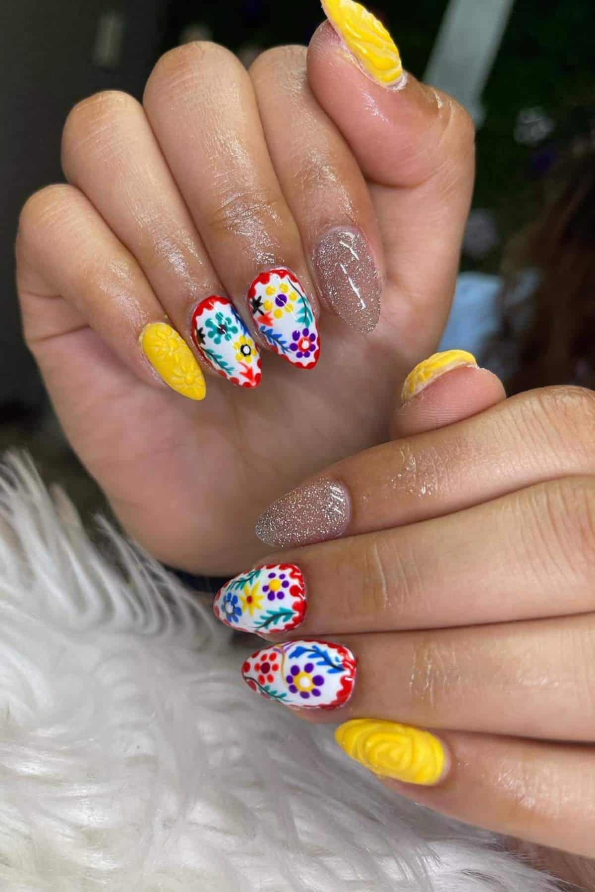 A hand with manicured nails featuring bright yellow, glittery nude, and white nails with colorful floral designs.