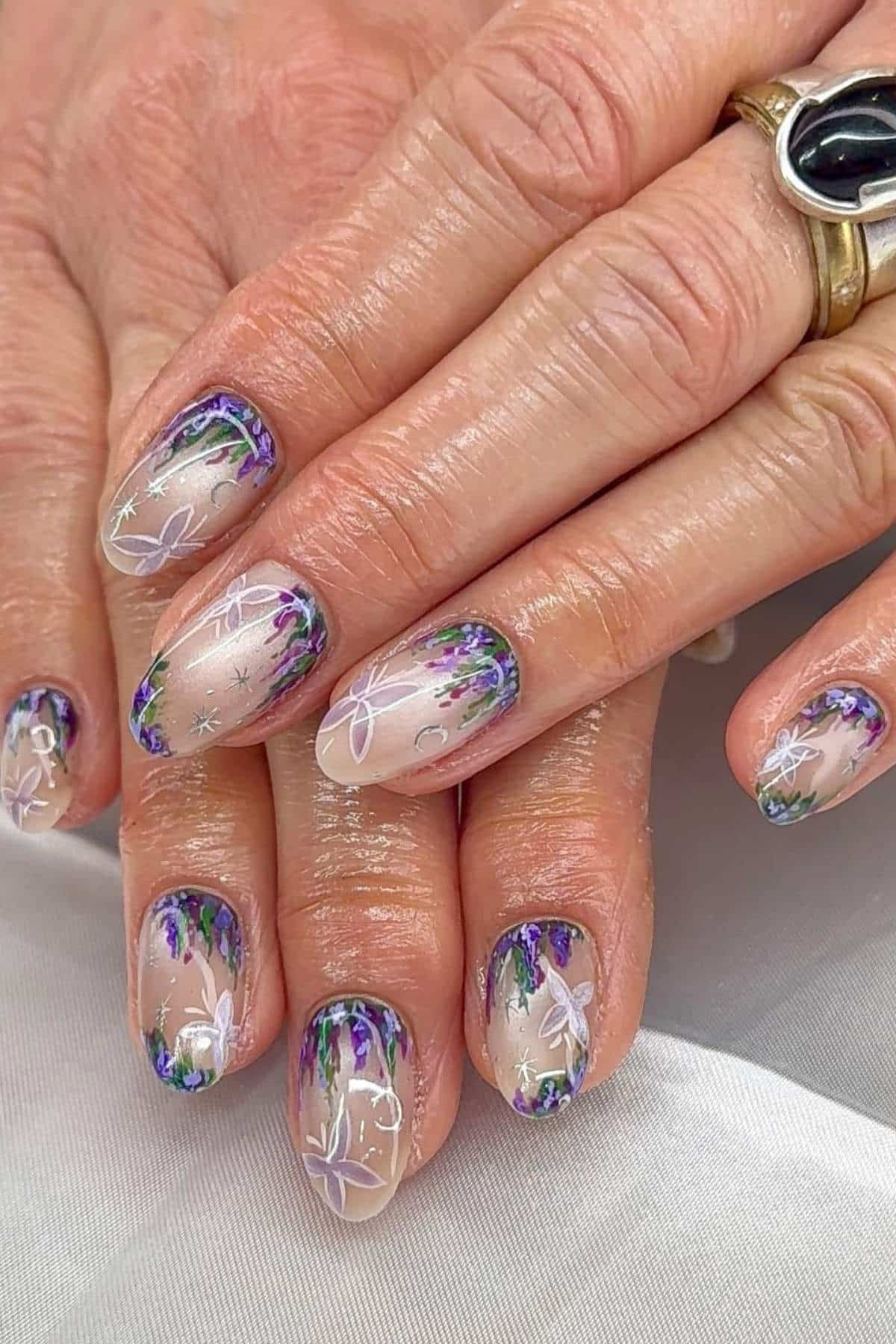 Hands with manicured nails featuring a clear base, purple and green floral designs, and white line art flowers.