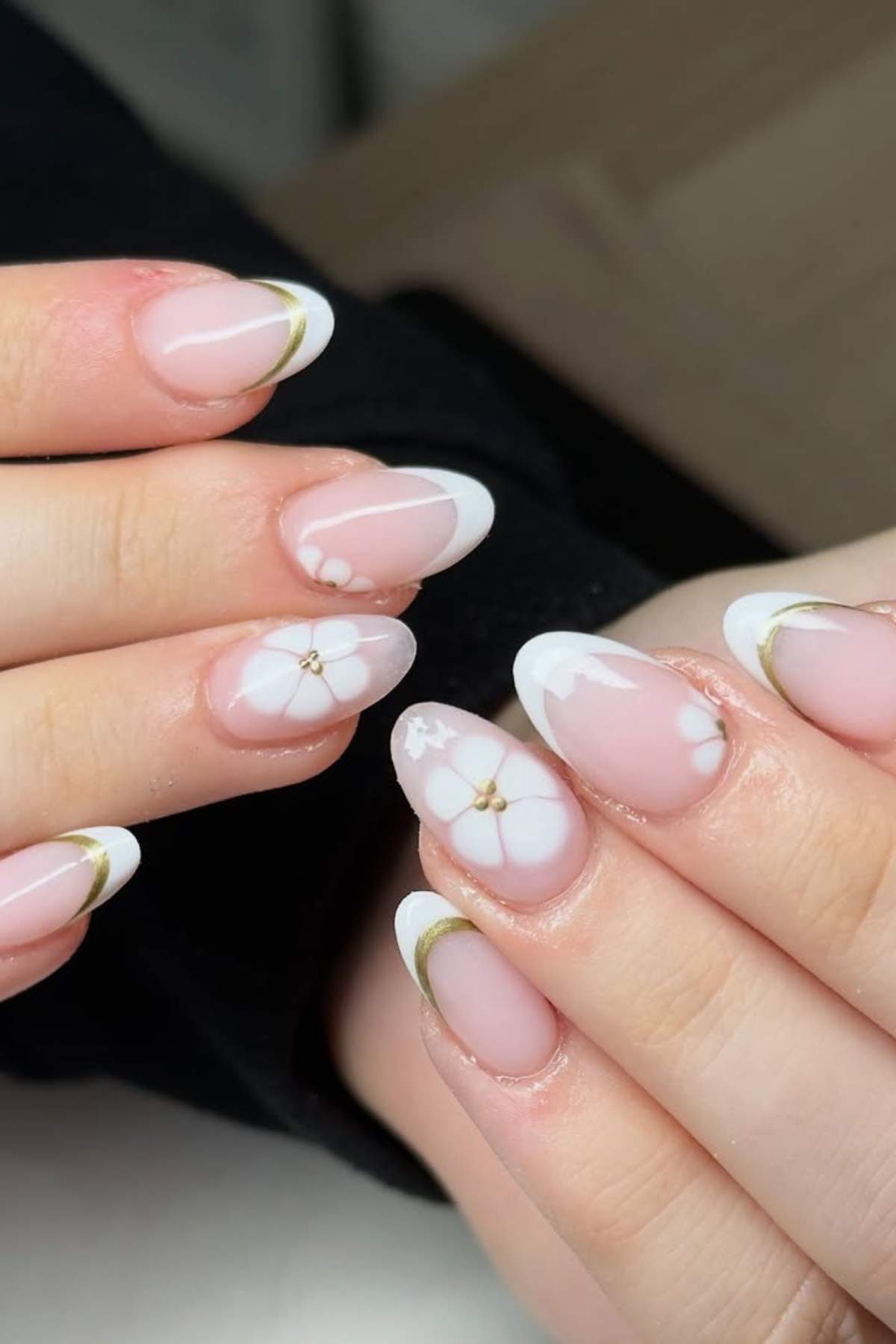 Hands with almond-shaped nails featuring a French tip design, white floral accents, and thin gold lines near the tips.