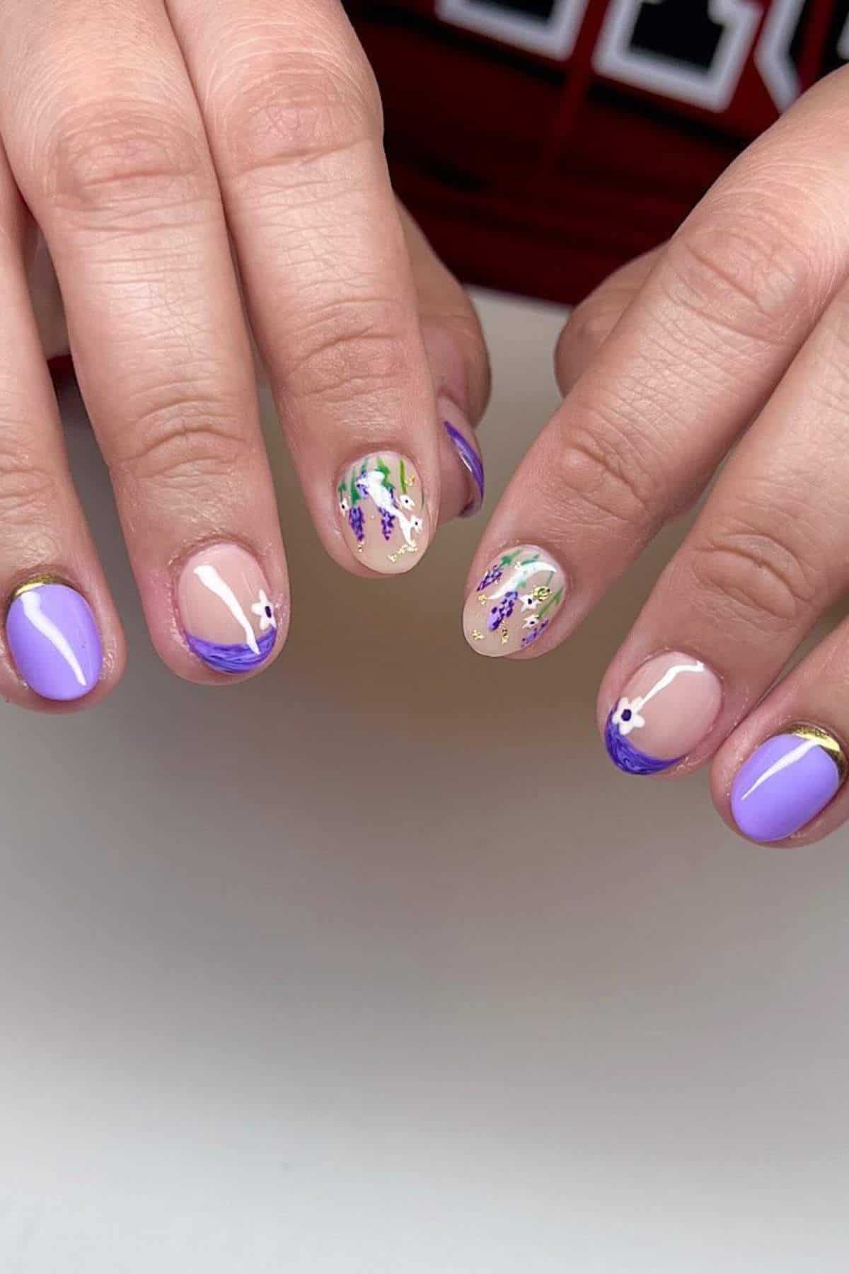 Hands with short, round nails featuring a mix of lilac polish, nude base, and floral designs in purple, green, and white on some nails.