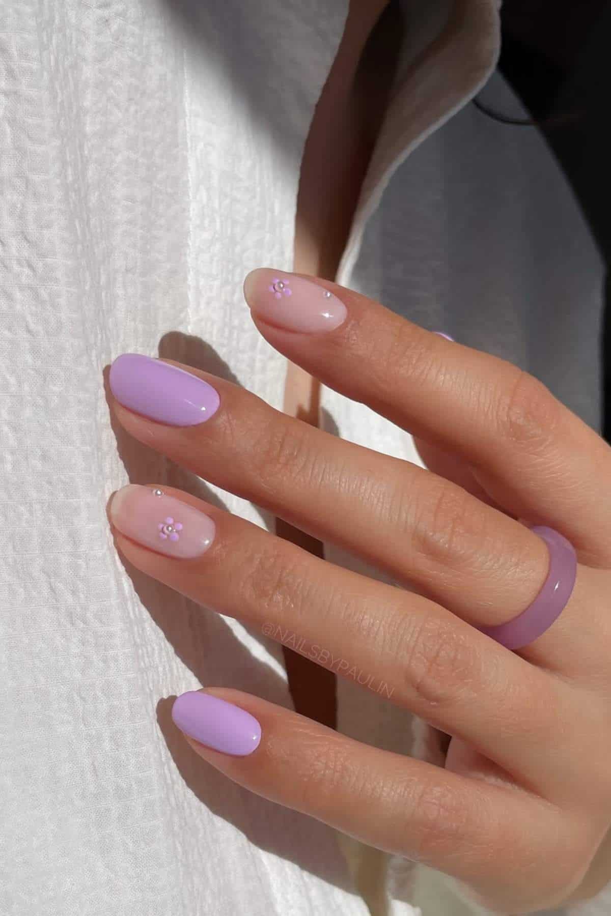 A hand with light purple and nude nails, some featuring small flower designs, rests against a white textured fabric. The person wears a matching purple ring.