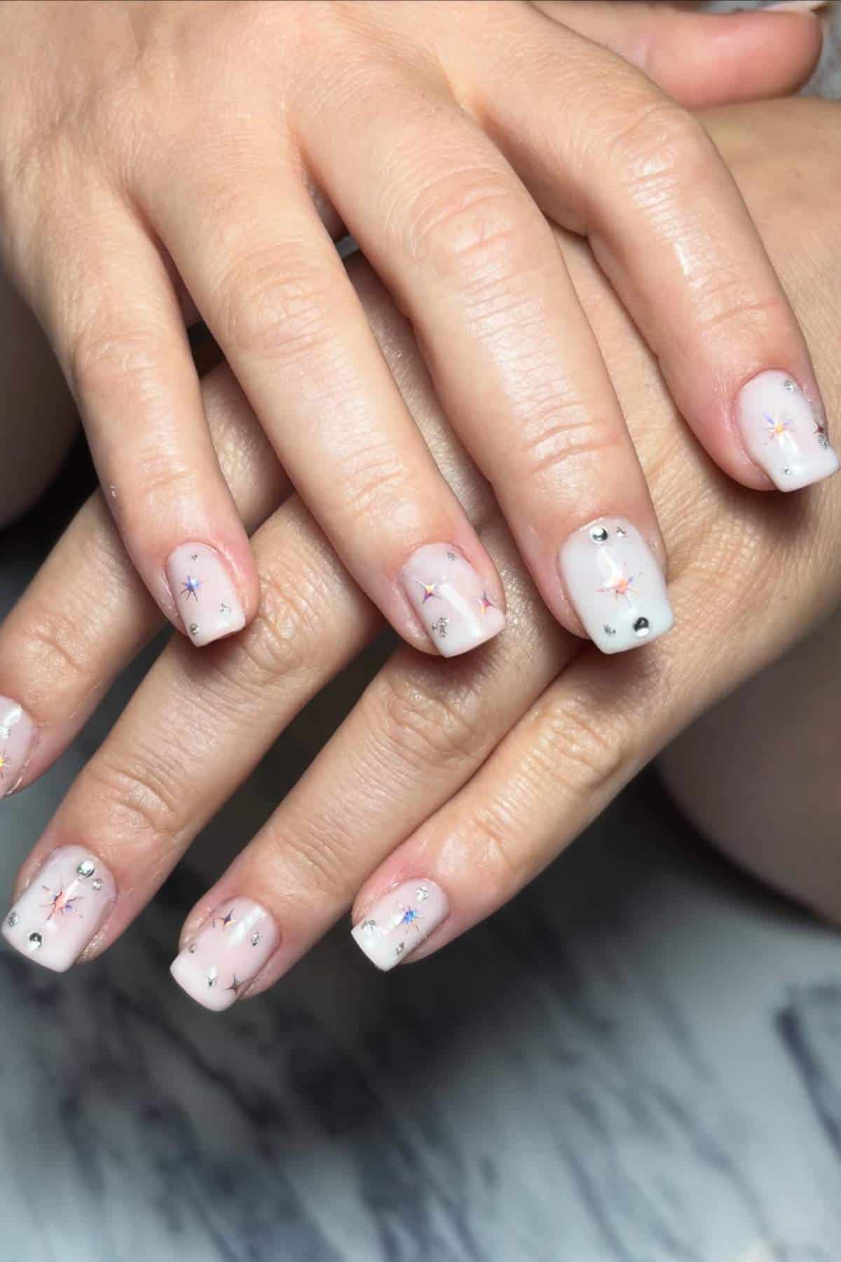 Hands with short, square-shaped nails painted white and decorated with small star designs and rhinestone accents, resting on a marble surface.