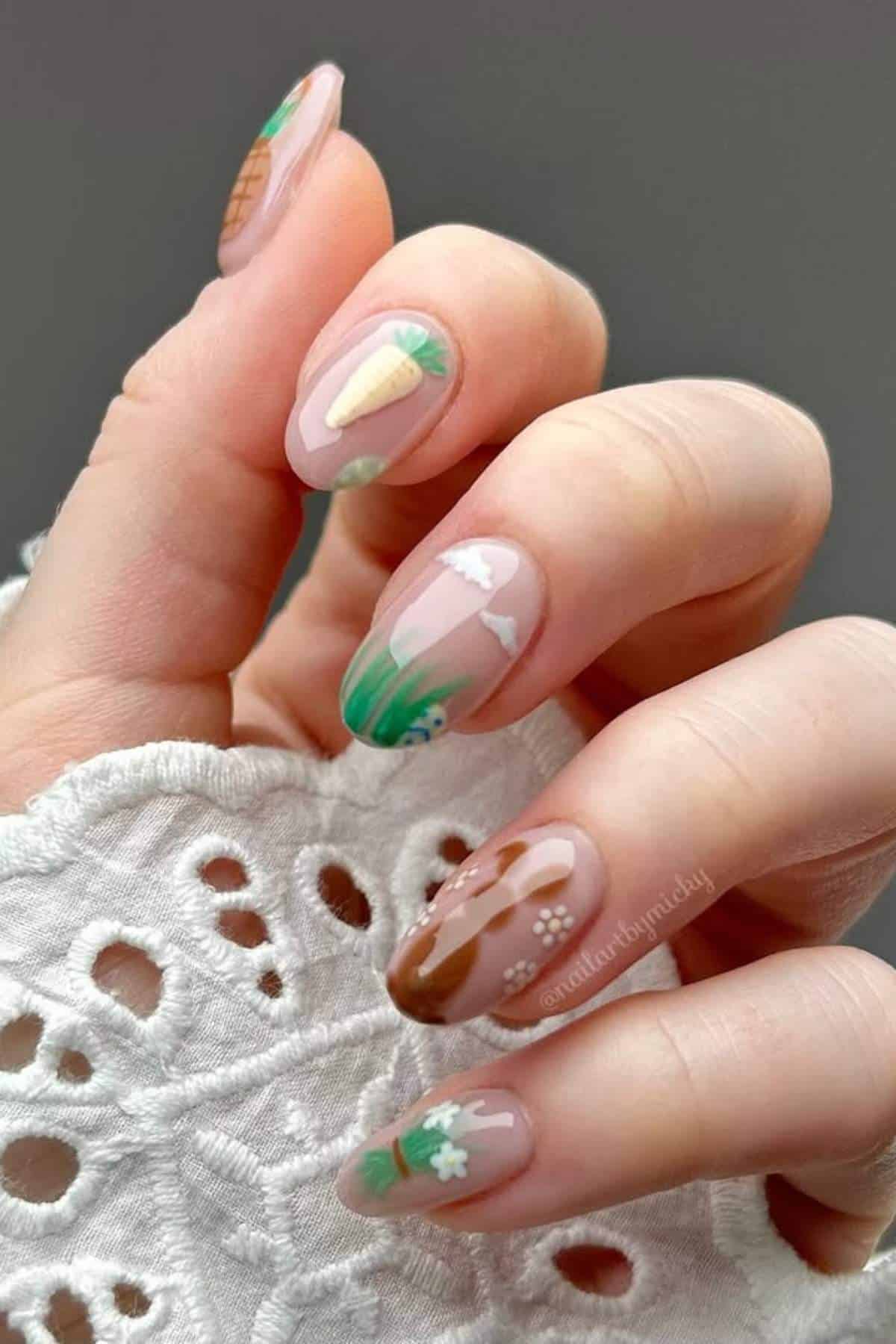 A hand with nude and green-themed nail art featuring designs of vegetables, plants, and flowers. The nails are medium-length and almond-shaped.