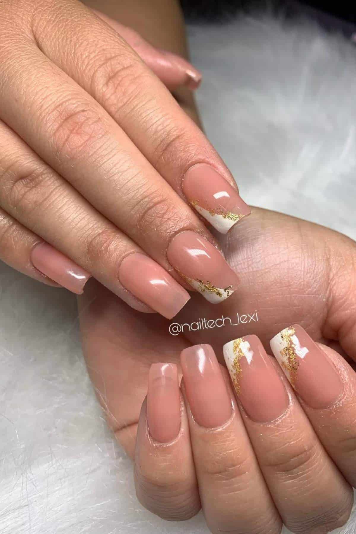 Hands with square-shaped, nude-toned acrylic nails featuring white tips and gold foil accents on some of the nails, displayed on a white surface.