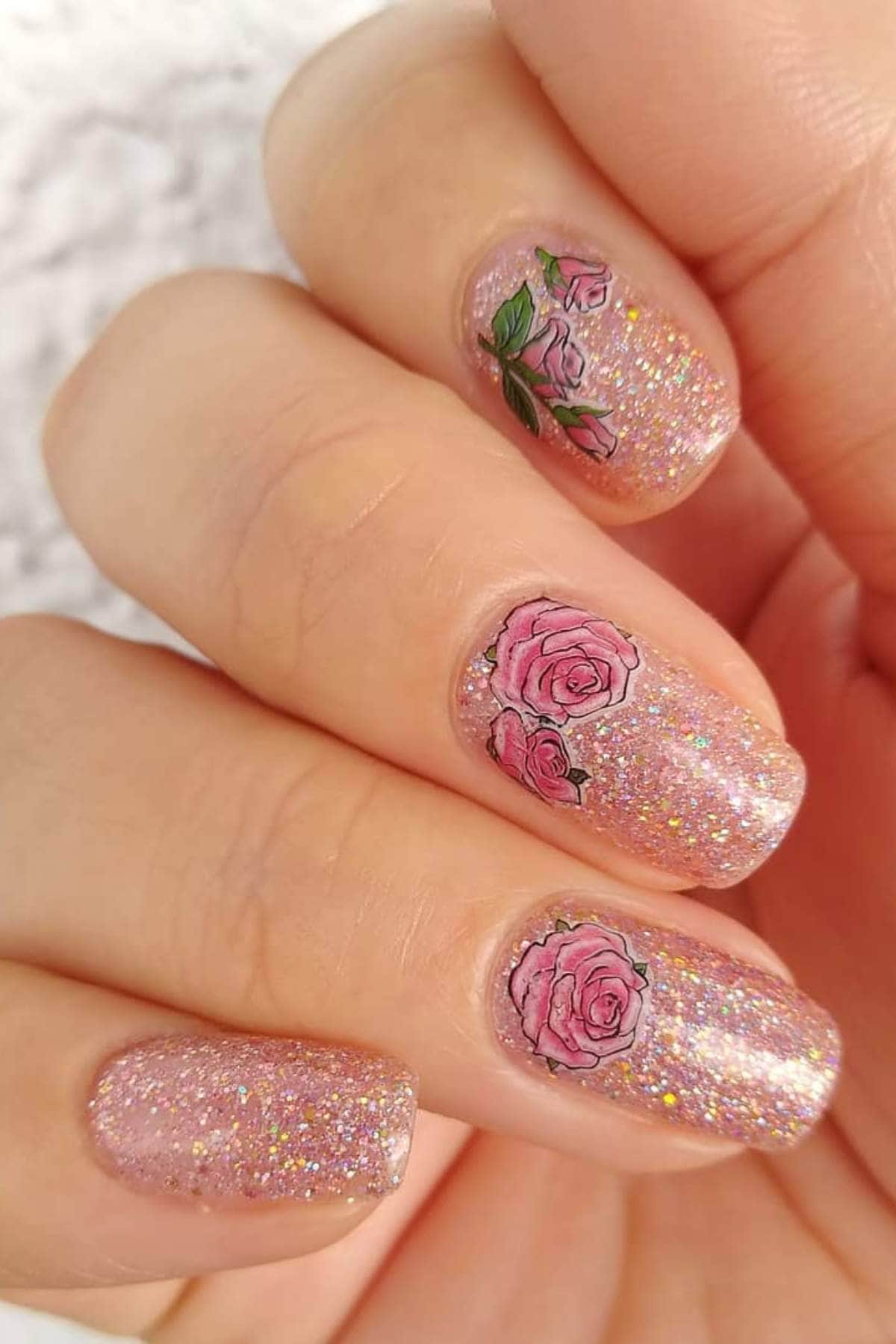 A hand with glittery pink nail polish features pink rose designs on three fingernails.