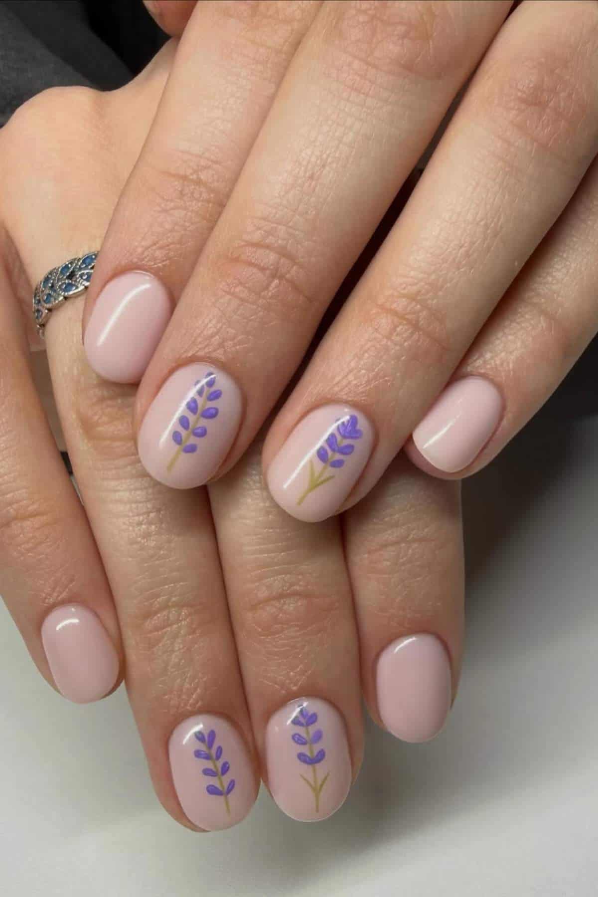 A hand with pale pink nails, two featuring purple and green lavender flower designs; person wears a silver ring on one finger.