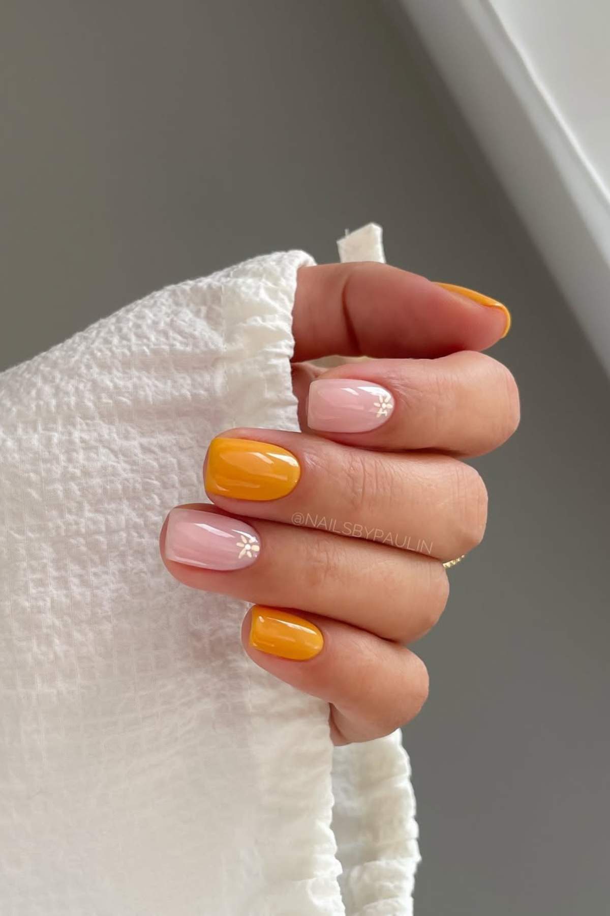 A hand with short nails painted in alternating yellow and light pink polish, with small white floral designs on two pink nails, holding a textured white fabric.
