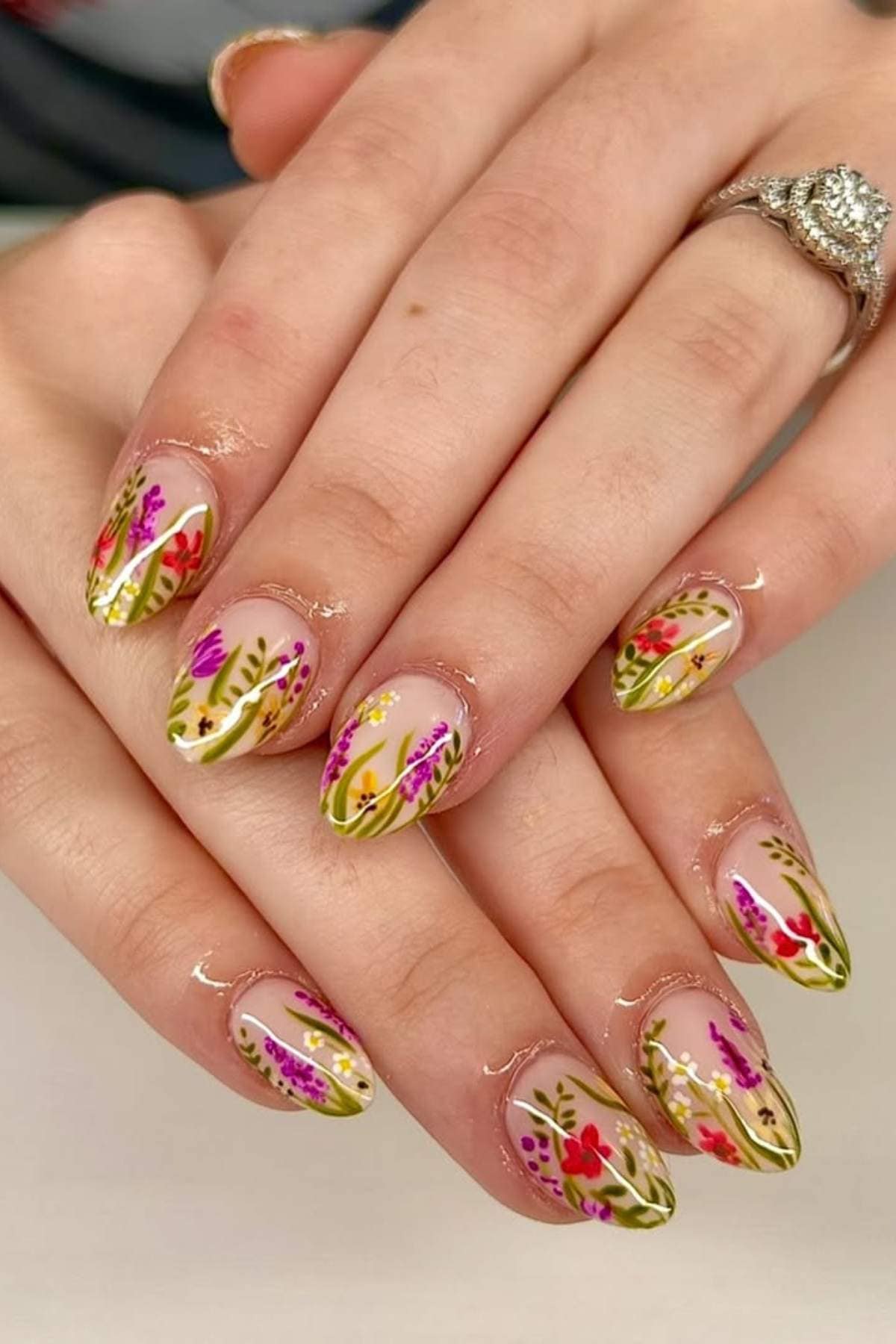 Hands with almond-shaped nails featuring a clear base and detailed floral designs in pink, red, and green. A silver ring with a central gemstone is on one finger.
