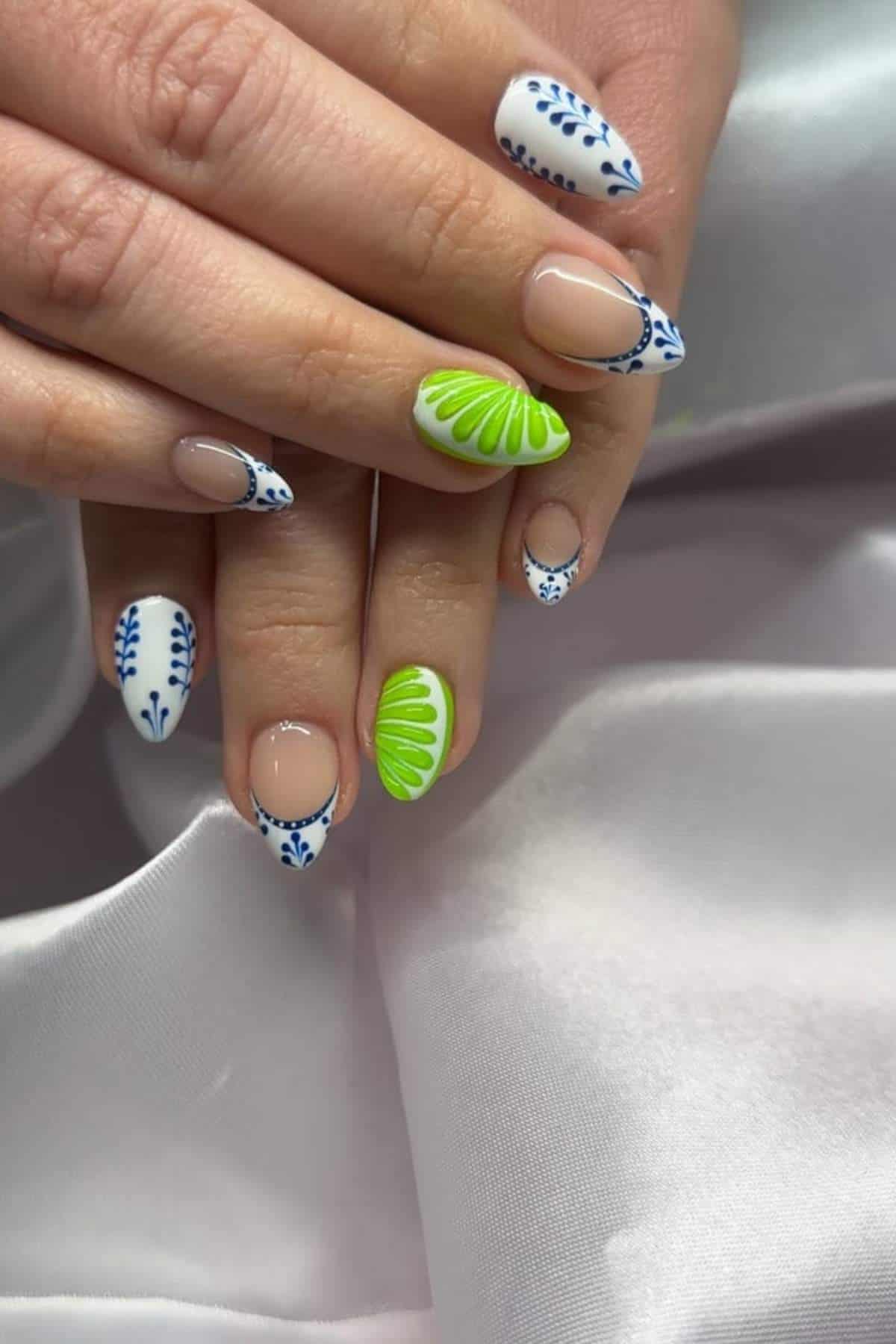 Close-up of hands with manicured nails featuring blue and white floral designs and bright green shell-like patterns on a light, satin fabric background.