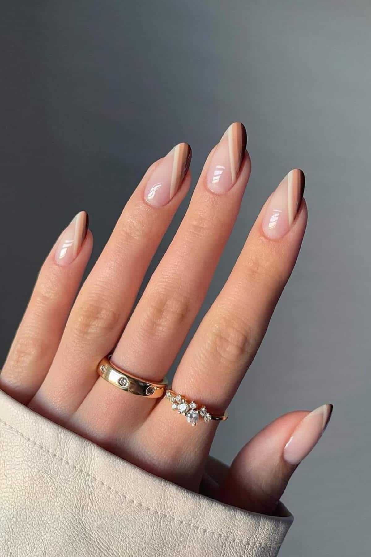 A hand with almond-shaped nails featuring a minimalist brown and nude diagonal stripe design, wearing two gold rings, one plain and one with small stones.