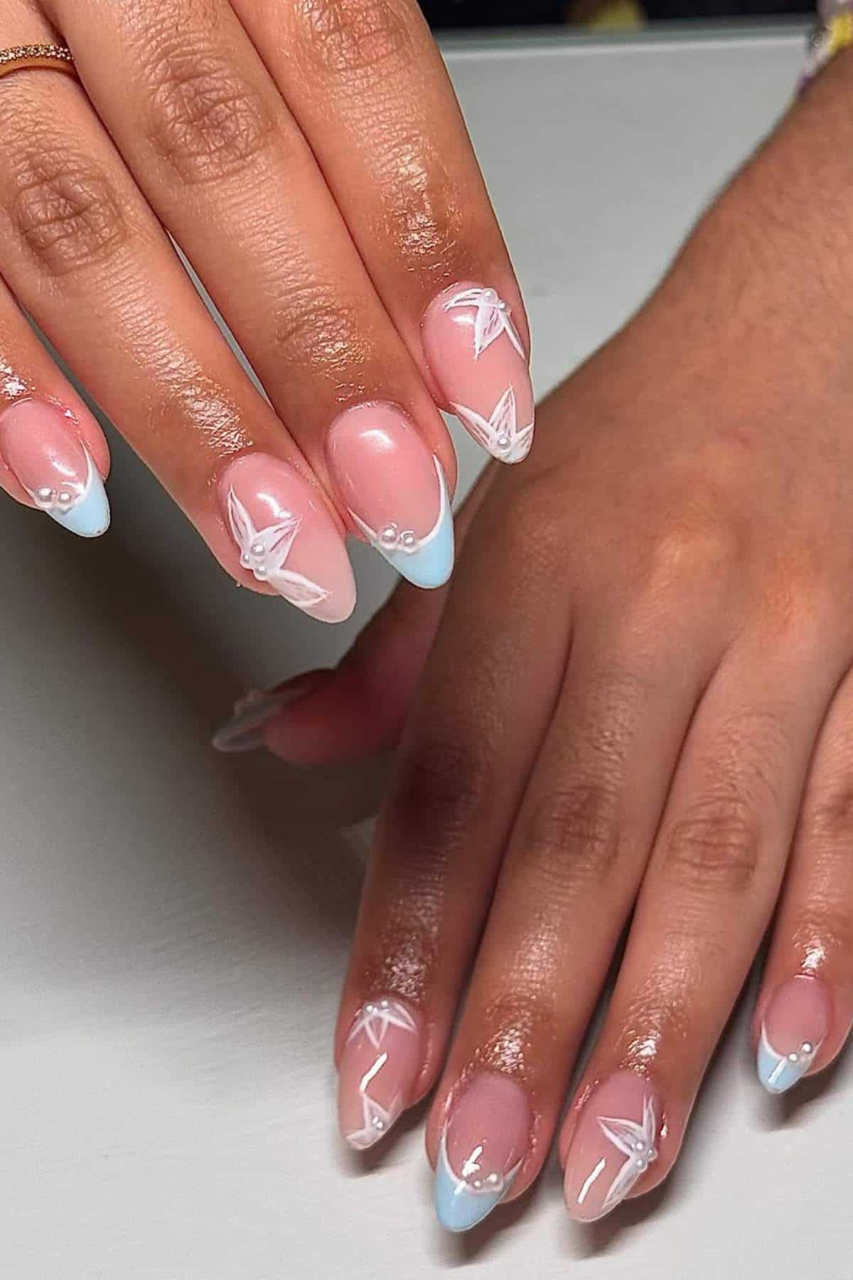 Hands with almond-shaped nails featuring a pink base, light blue French tips, and white floral designs with small bead accents.