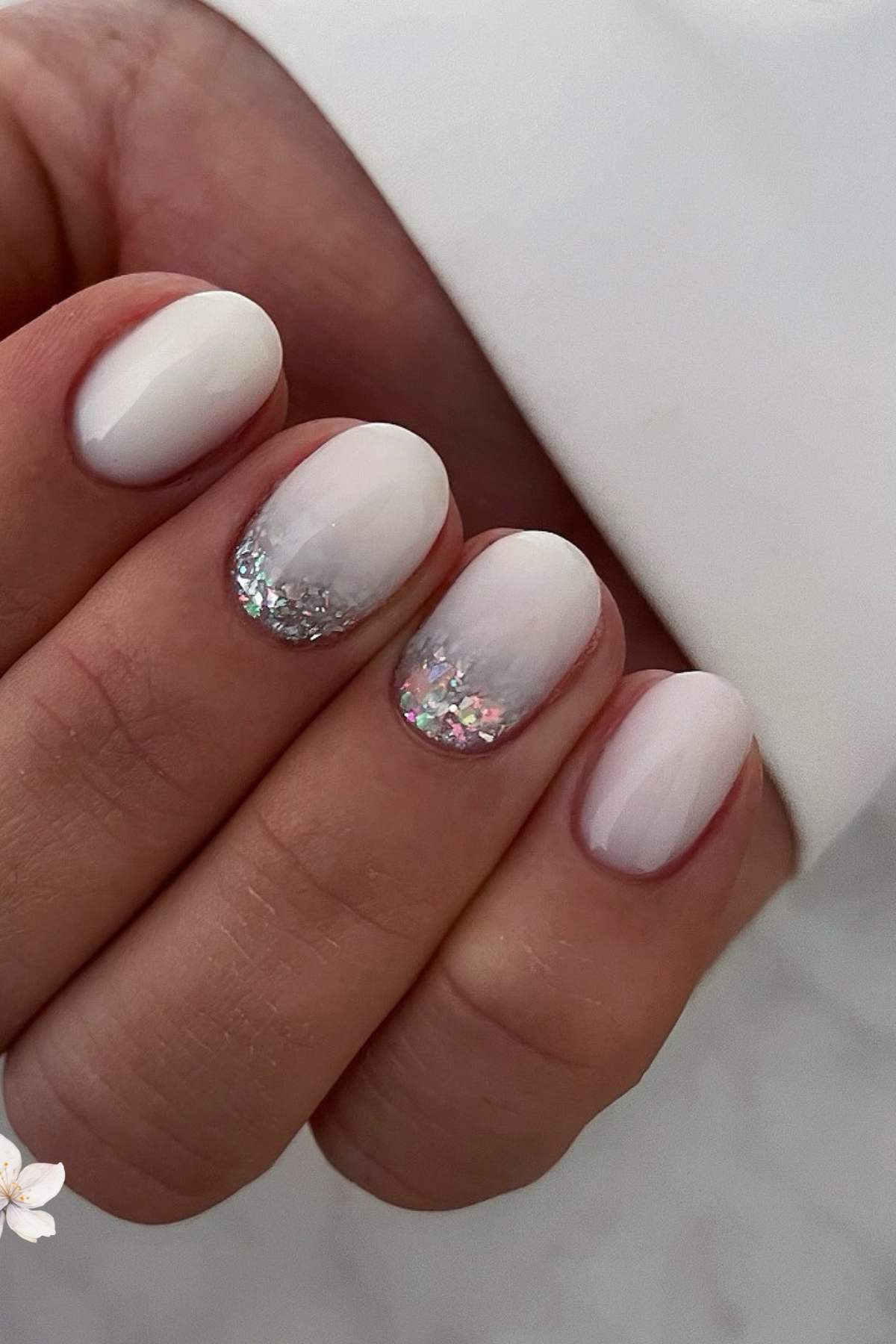 A hand with short, oval-shaped nails painted white, featuring silver glitter accents on the ring and middle fingernails.