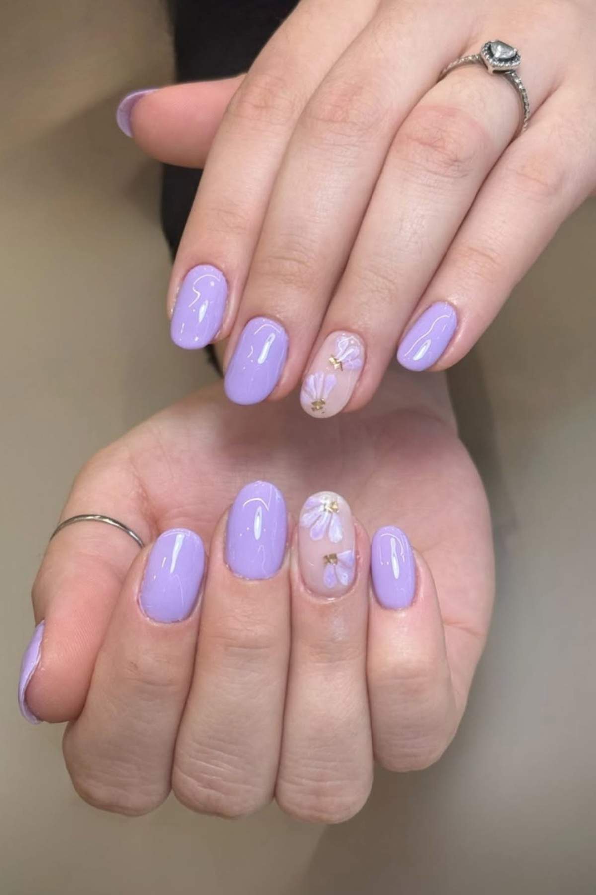 Hands with short, oval-shaped nails painted light purple; ring fingers feature clear polish with small white and yellow floral designs.