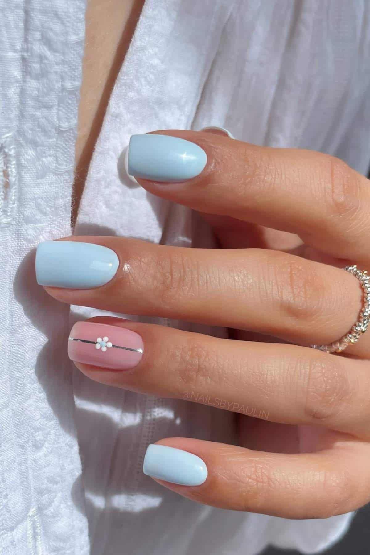 A hand with light blue square-shaped nails; the ring fingernail is pink with a white flower and black lines. The person wears a textured white shirt and a silver ring.