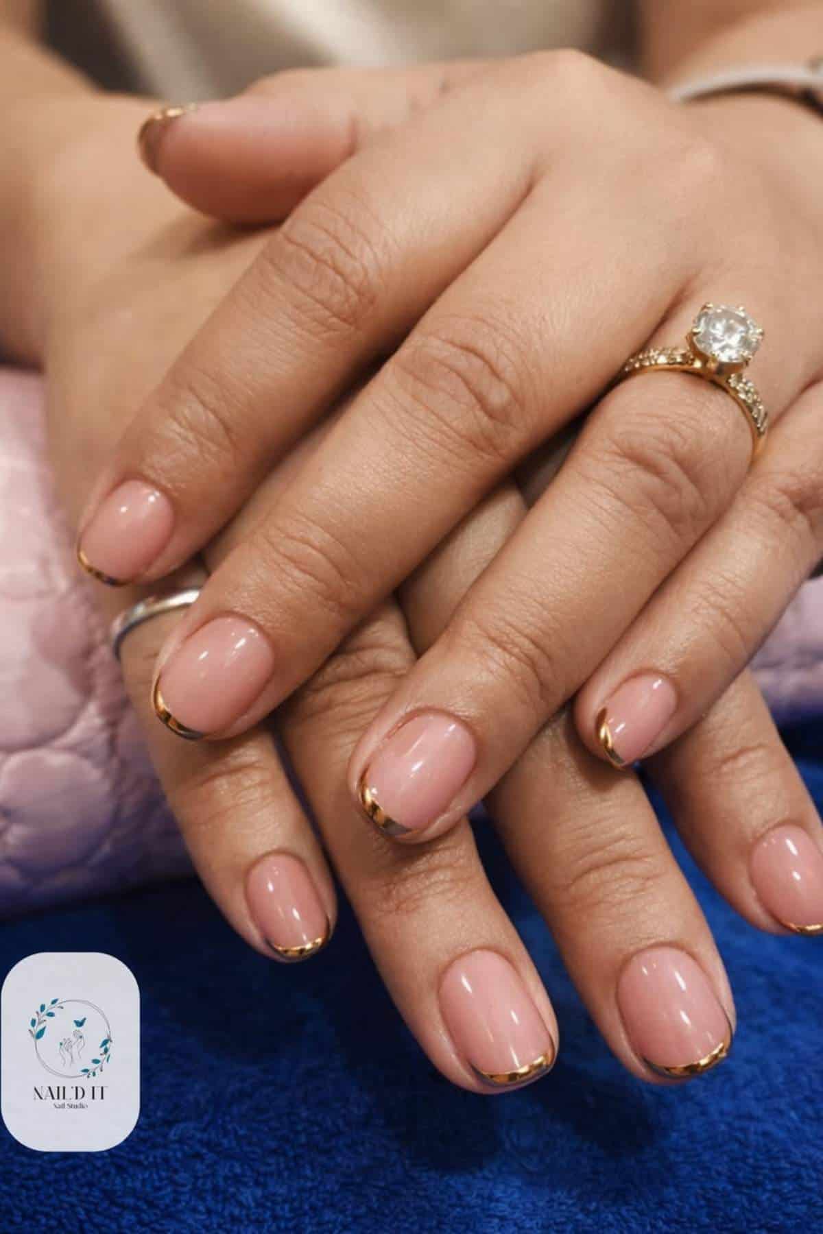 Close-up of hands with short, natural-shaped nails featuring a nude base and thin gold French tips; person wears a gold ring with a large clear stone.