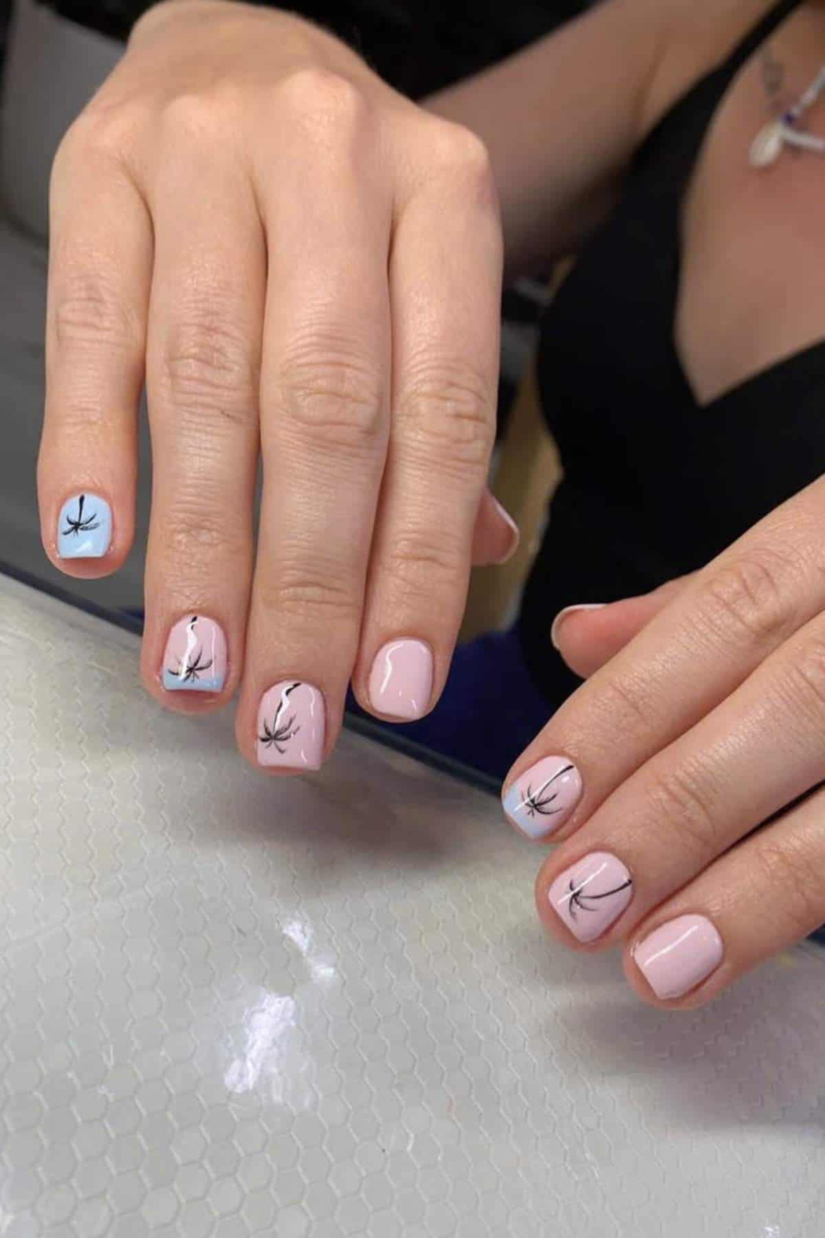 A person’s hands with short, light pink nails featuring black abstract branch designs and light blue accents on some nails, resting on a textured white surface.