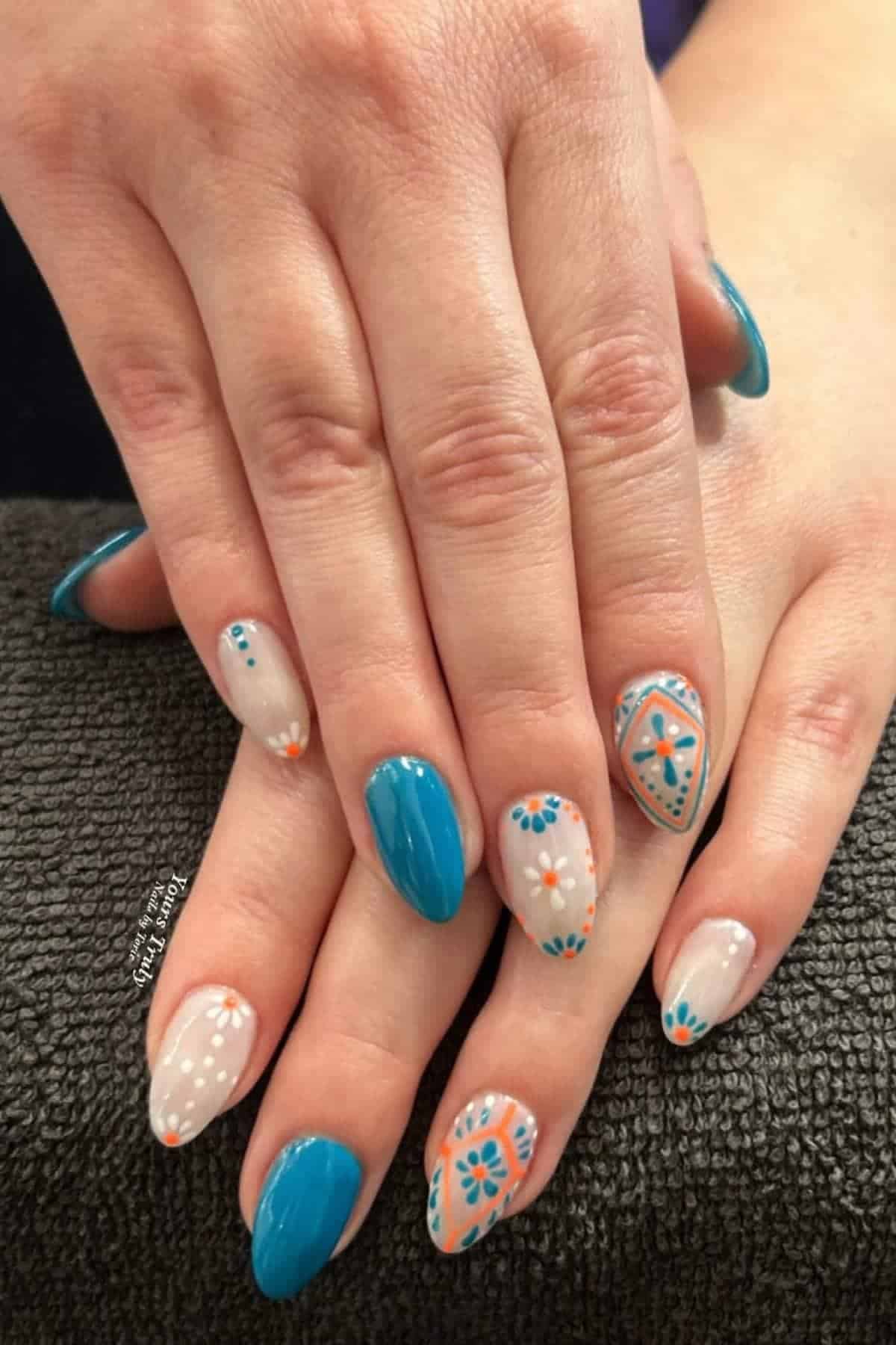 A pair of hands with manicured nails featuring blue, white, and orange floral and geometric designs on some nails, and solid blue polish on others, resting on a dark textured surface.