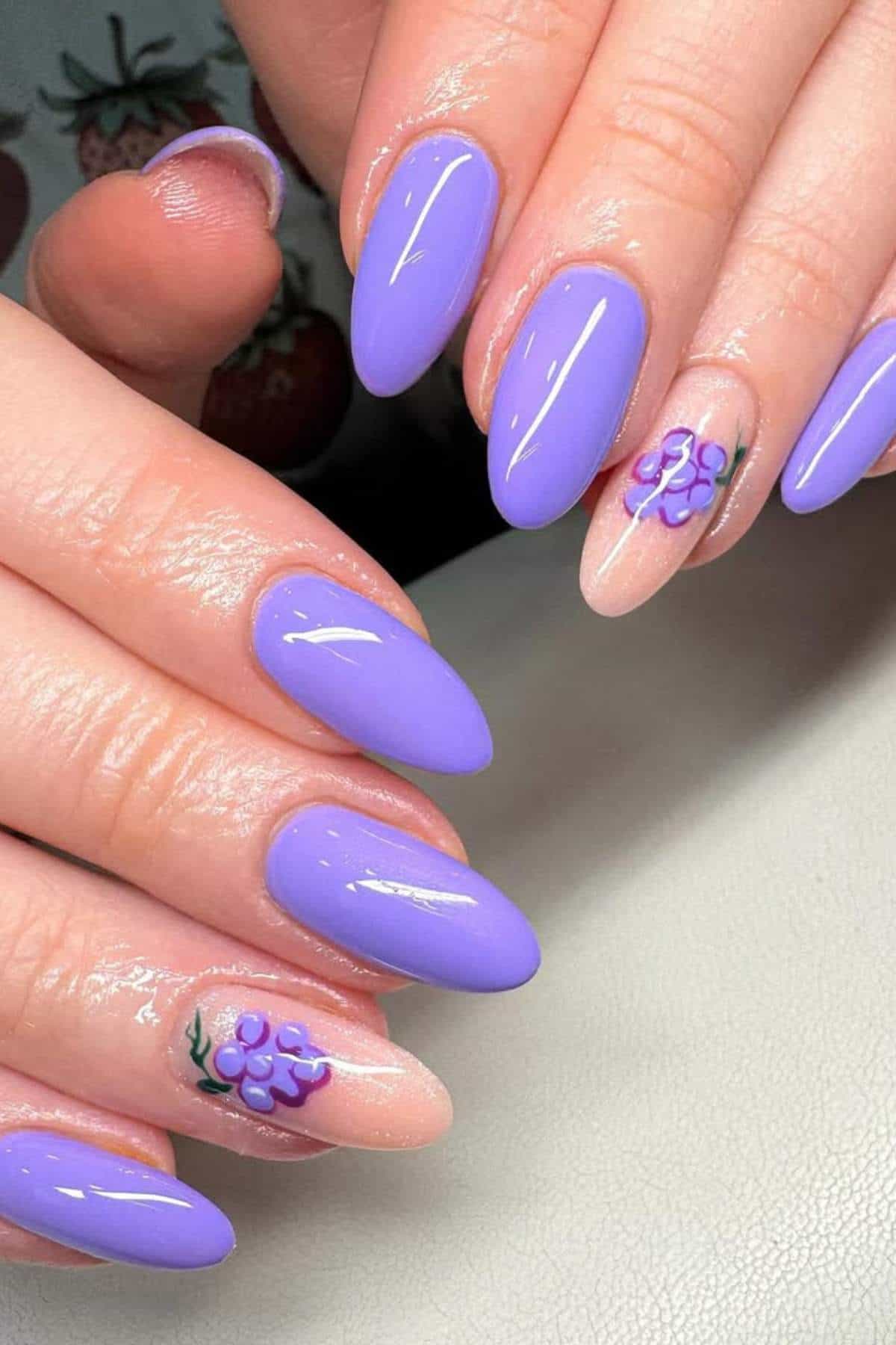 Hands with almond-shaped nails painted in glossy lavender polish, with two accent nails featuring small purple flower designs on a nude base.