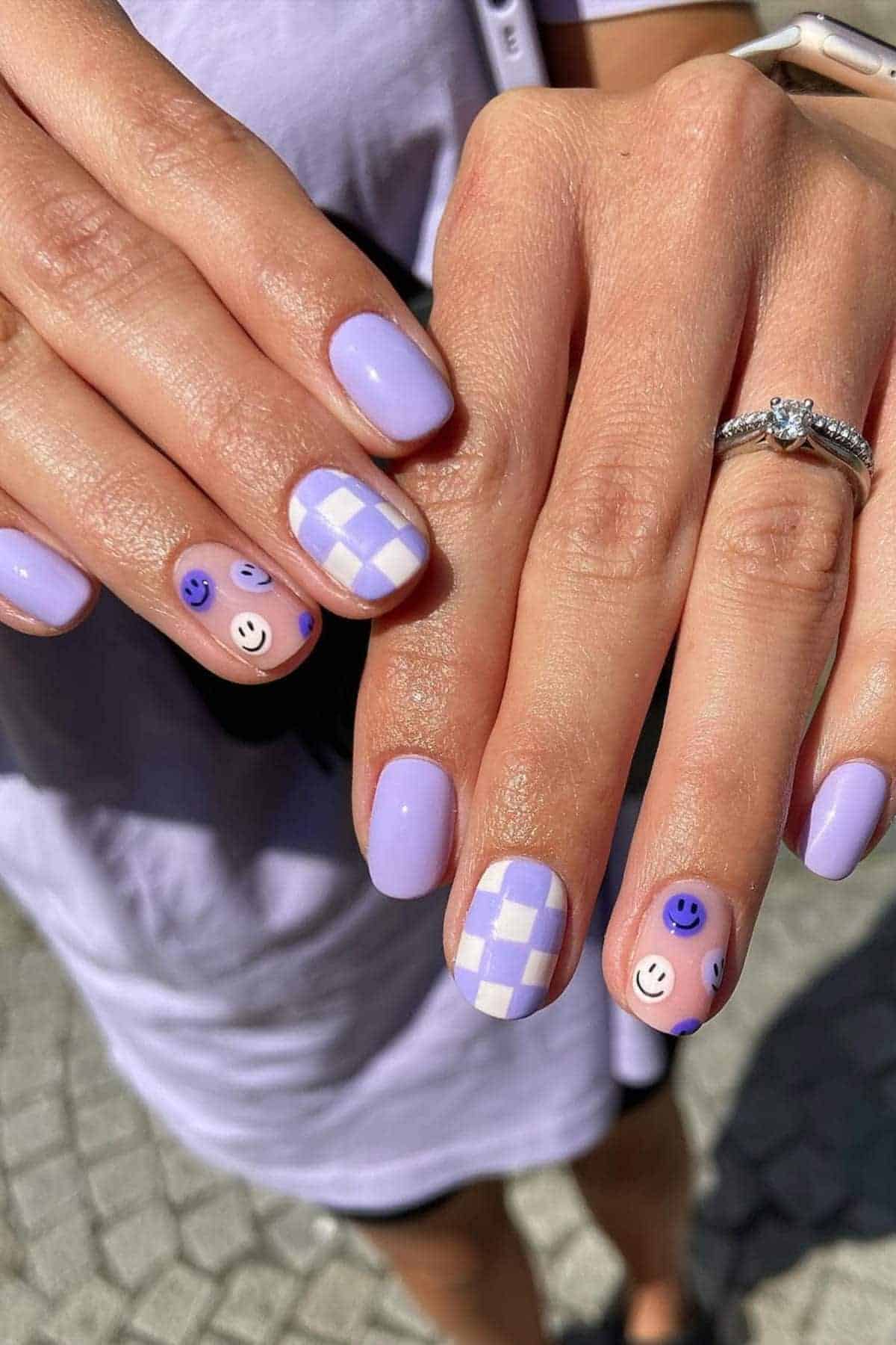 Hands with light purple manicure, featuring checkered and smiley face designs on accent nails. Person is wearing a silver ring and a smartwatch.