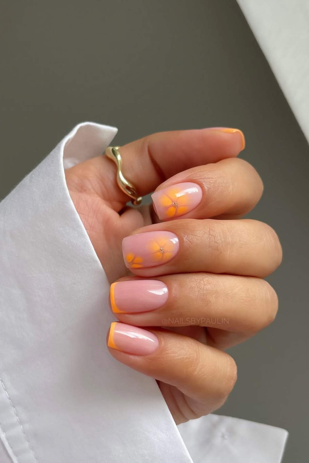 A hand with a gold ring displaying manicured nails with a light pink base, orange French tips, and yellow flower designs on two nails.