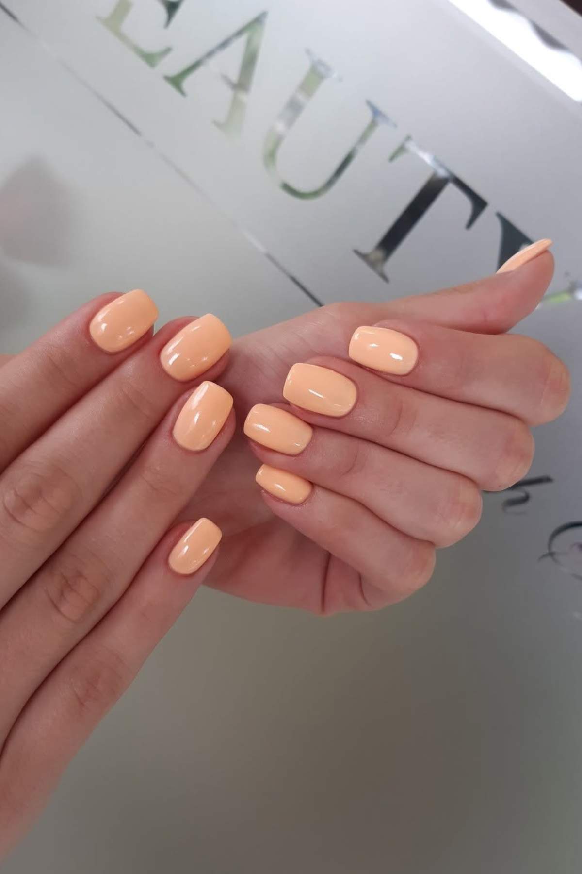 Close-up of hands with neatly manicured nails painted in a glossy peach color, held in front of a blurred background with partially visible text.