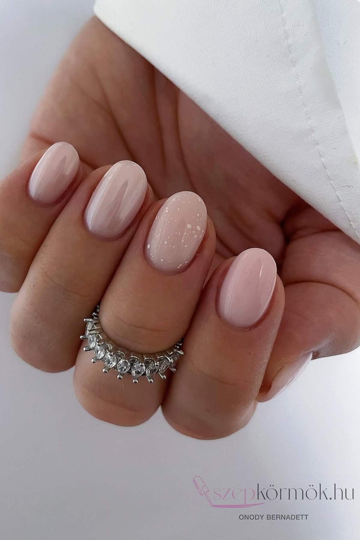 A hand with short, oval-shaped nails painted in glossy light pink polish; one nail has a subtle white speckled design. The person is wearing a jeweled ring.
