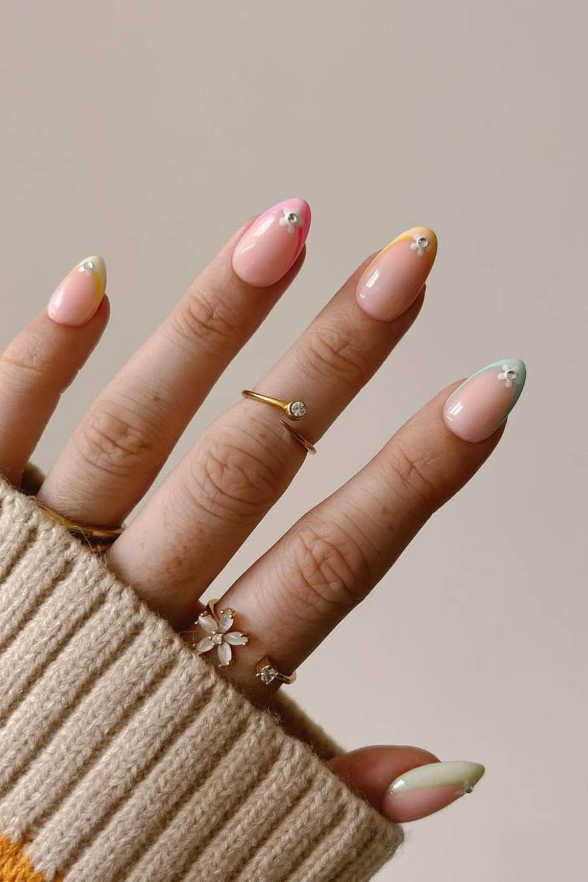 A hand with pastel-colored French tip nails, each topped with a small white flower design. The person is wearing gold rings and a beige sweater.