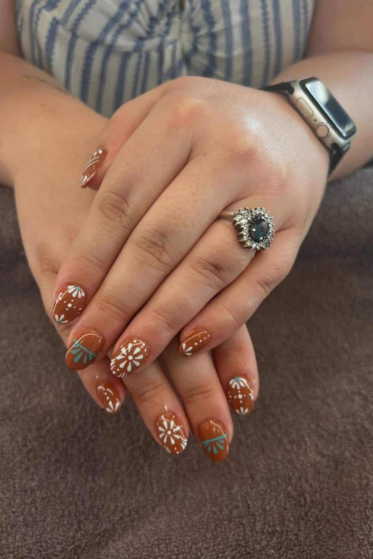 Hands with brown nail polish featuring white and green floral designs, wearing a silver ring with a dark stone, resting on a brown towel.