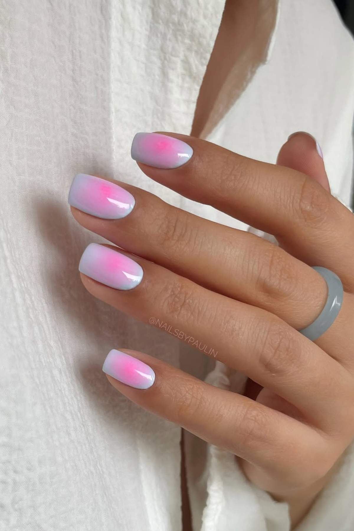 A hand with short, square-shaped nails featuring a soft gradient pink and white ombre manicure, resting on a white textured fabric.