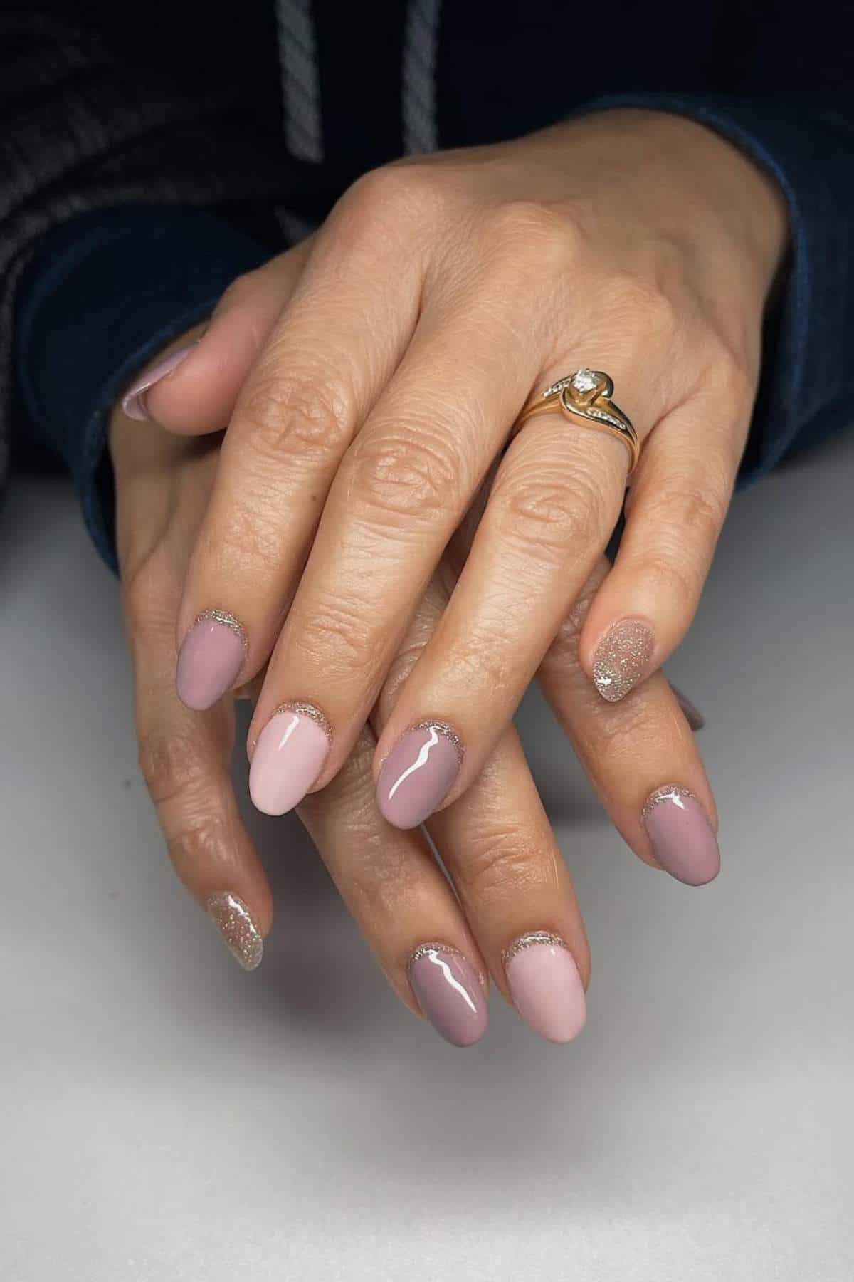 Hands with almond-shaped nails painted in shades of pink and mauve, accented with glitter near the cuticles. A gold ring with a clear stone is worn on the ring finger.