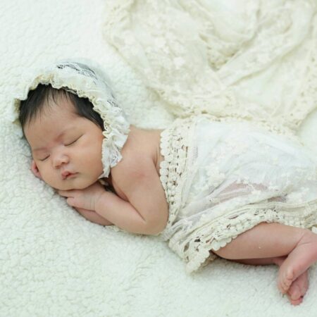 A newborn baby sleeps on a soft white blanket, dressed in a delicate lace bonnet and wrap.