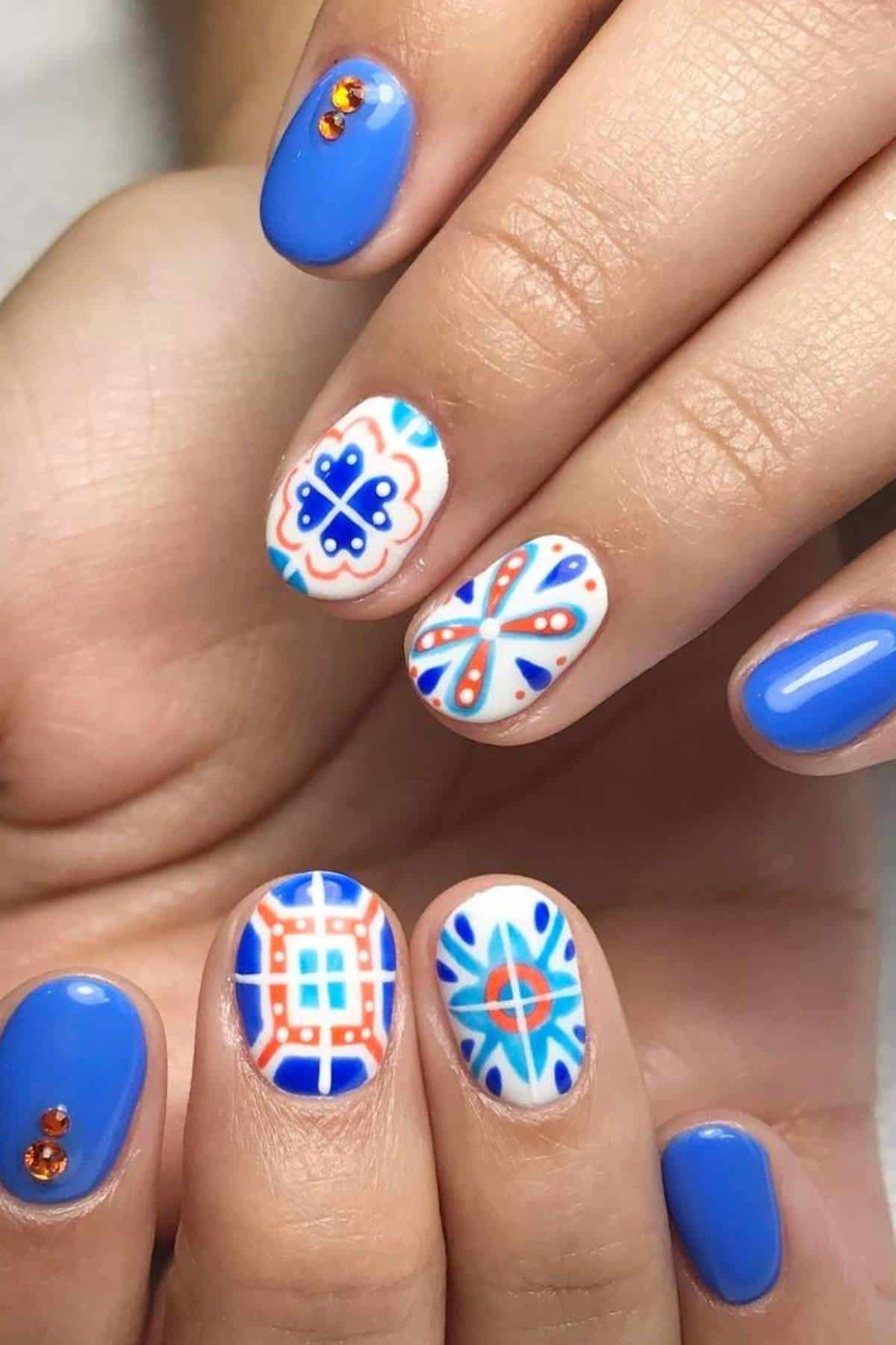 Hands with blue manicure; some nails feature intricate blue, orange, and white geometric designs, while others have solid blue polish with orange rhinestone accents.