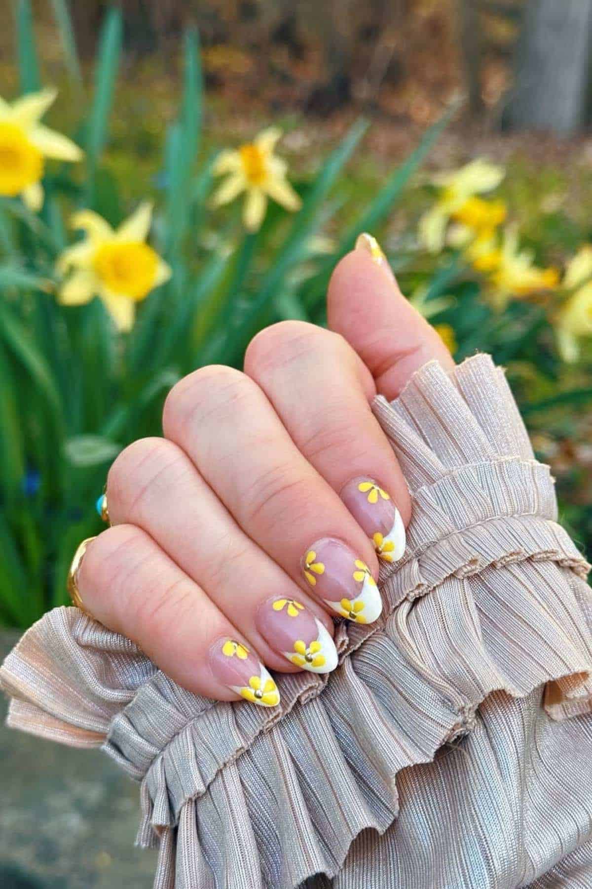 A hand with long nails painted with yellow and white floral designs is held up in front of blooming yellow flowers outdoors.