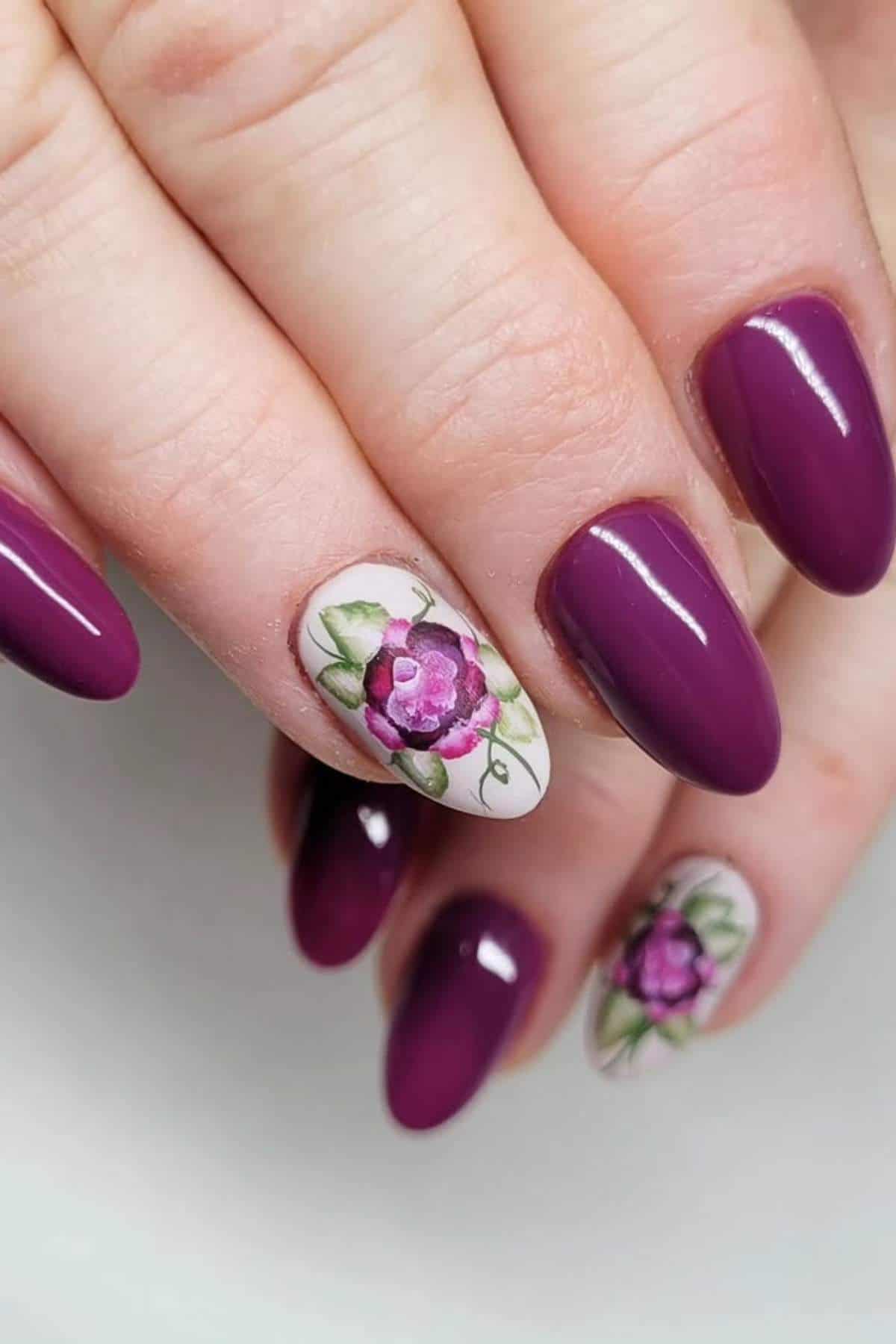 A hand with almond-shaped nails painted purple, with two accent nails featuring detailed floral designs in purple, green, and white.