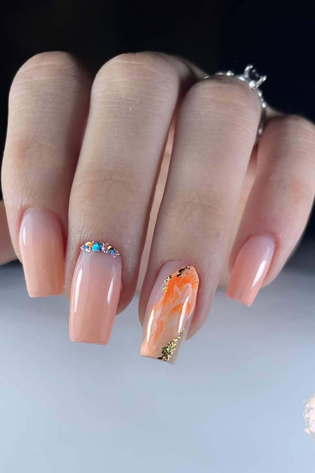 A hand with square-shaped nude nails; one nail has orange and gold marble art, another features small rhinestones near the cuticle.