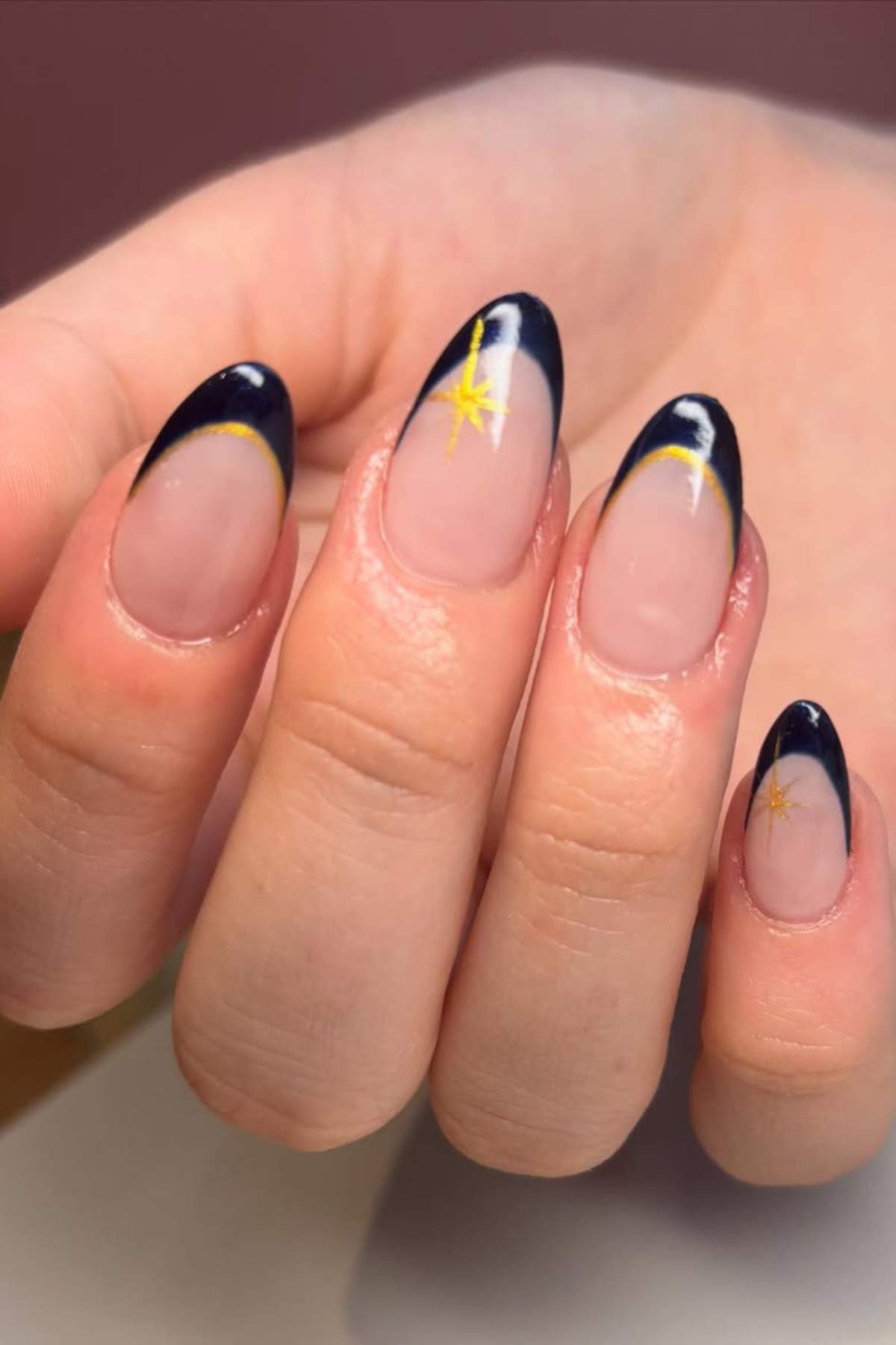 A hand with almond-shaped nails featuring a dark blue French tip and a small yellow star design on two nails.