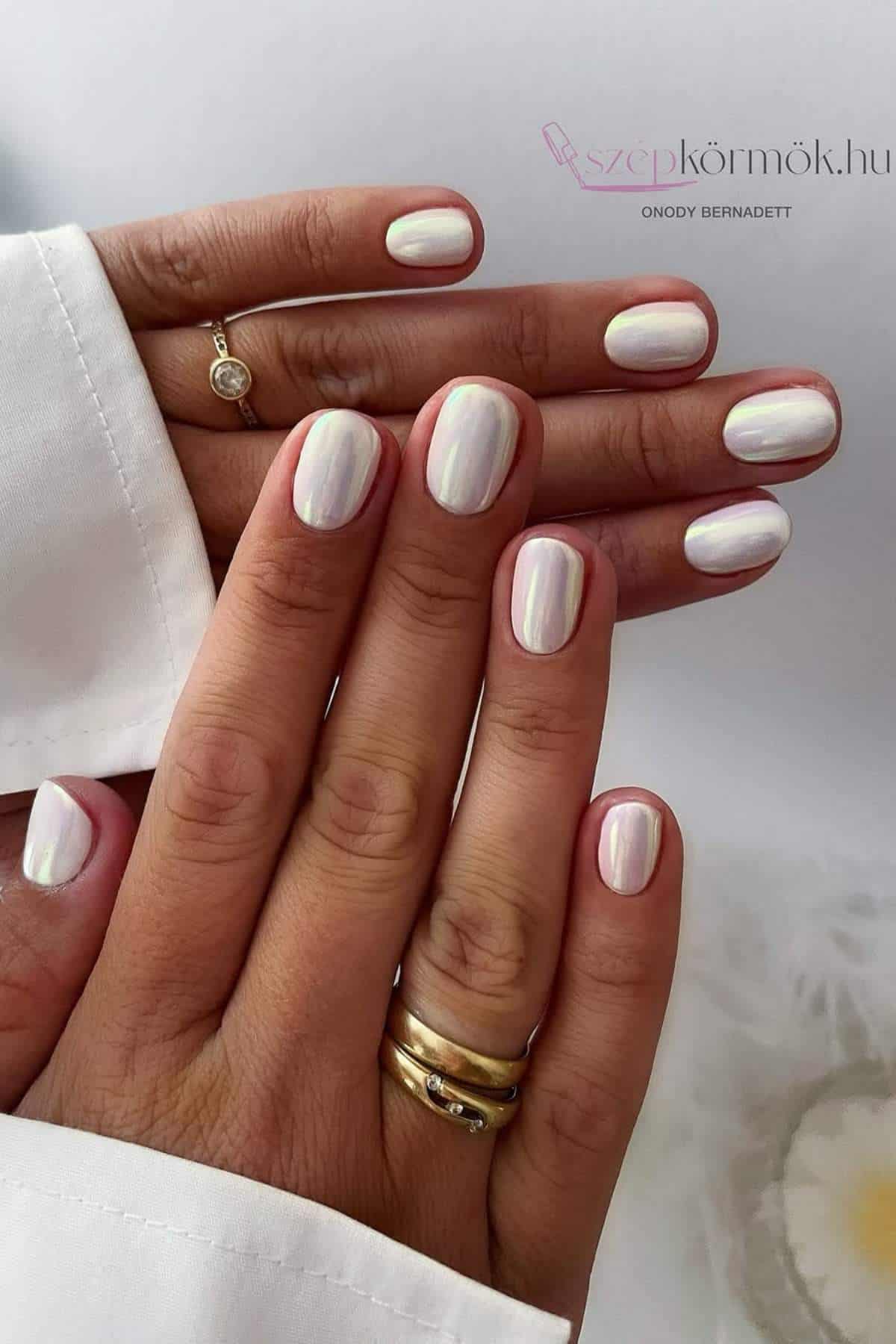 Close-up of hands with short, neatly manicured nails painted in a shiny, pearly white polish. The person is wearing gold rings and a white long-sleeve garment.