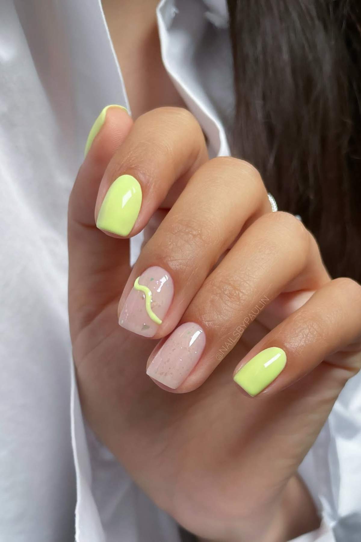 A hand with short, square-shaped nails painted in yellow and nude pink with a wavy yellow accent design on the ring finger.