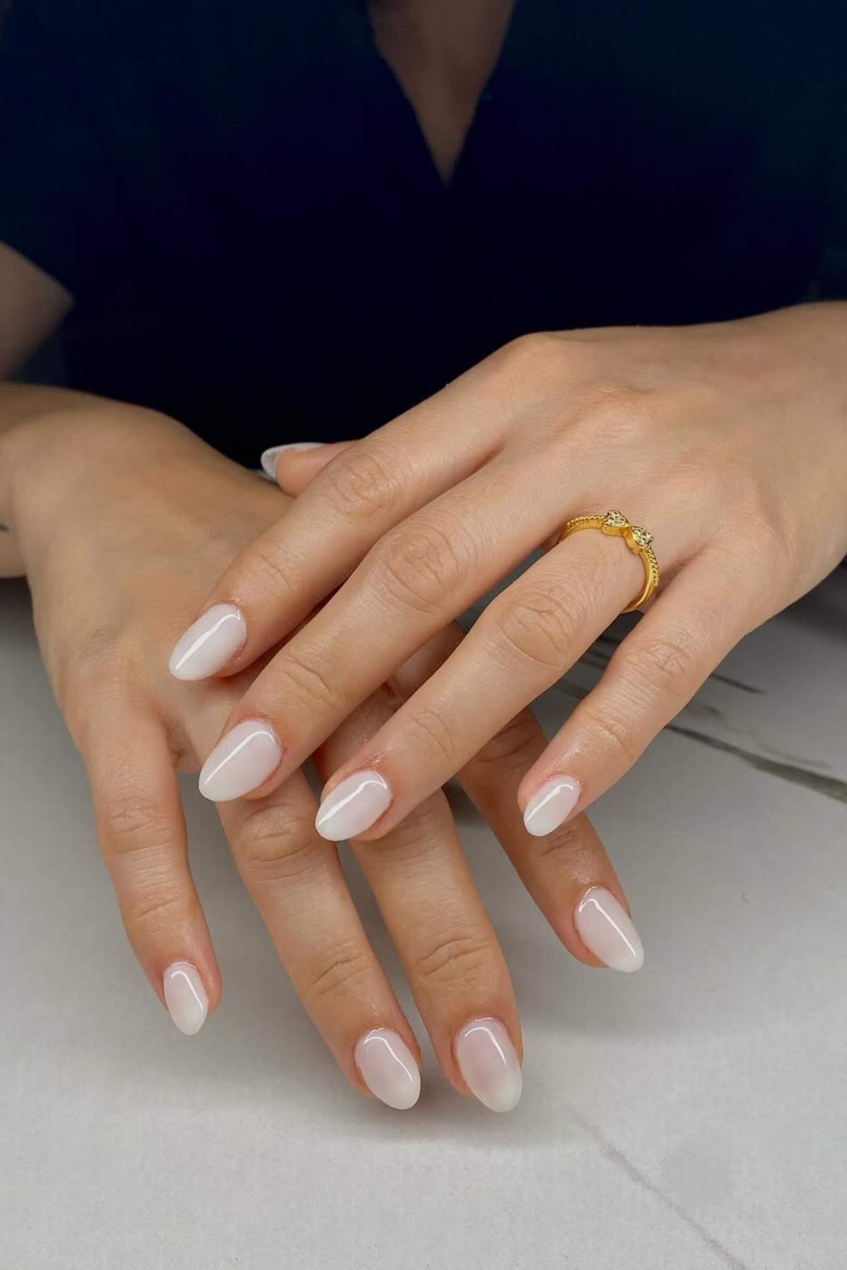 Hands with almond-shaped nails painted in a glossy pale pink shade, one hand wearing a gold ring with stones, resting on a white surface.
