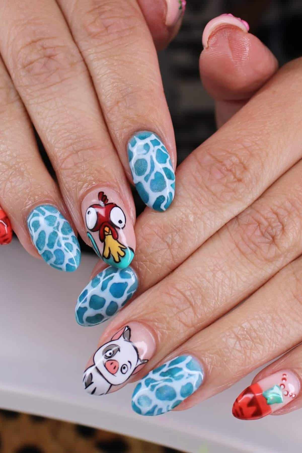 Close-up of hands with manicured nails featuring blue scales pattern, a cartoon chicken, and a cartoon pig design on the ring and pinky fingers.