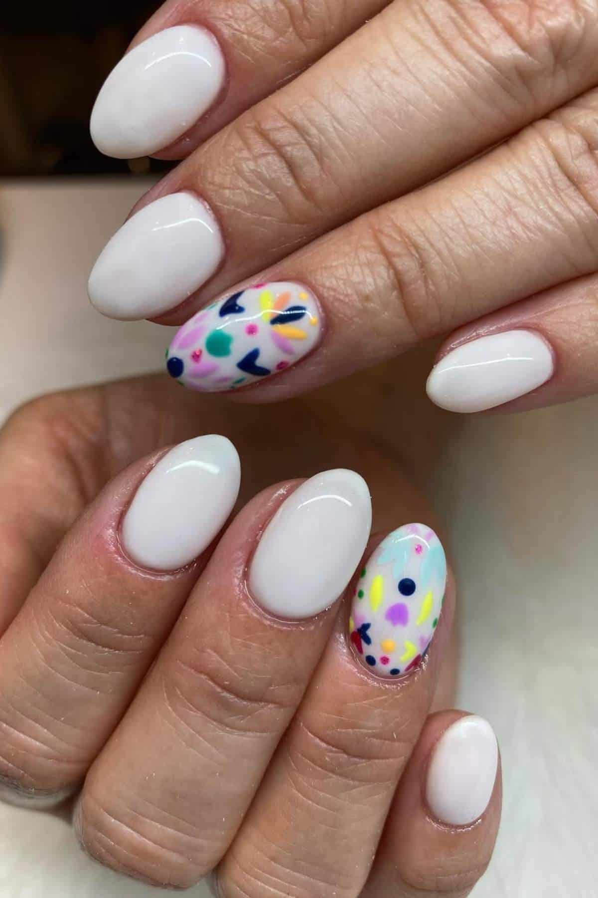 Hands with oval-shaped nails painted white, featuring colorful abstract designs on the ring fingers.