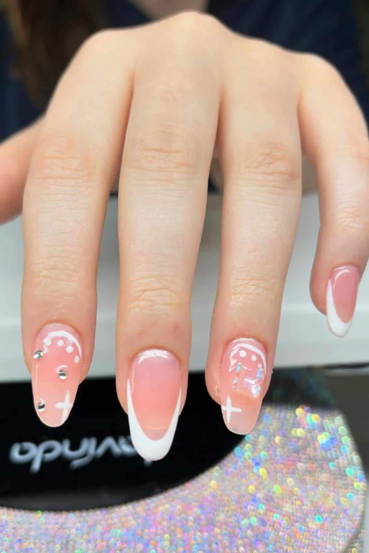 A hand with long, almond-shaped nails featuring a French manicure, white designs, and small silver rhinestones on two nails.