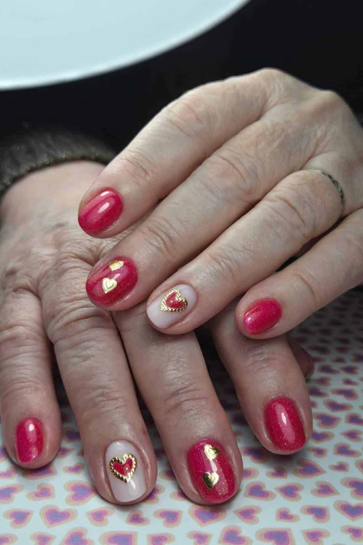 Close-up of hands with short, rounded red nails featuring gold and red heart-shaped nail art on a patterned surface.