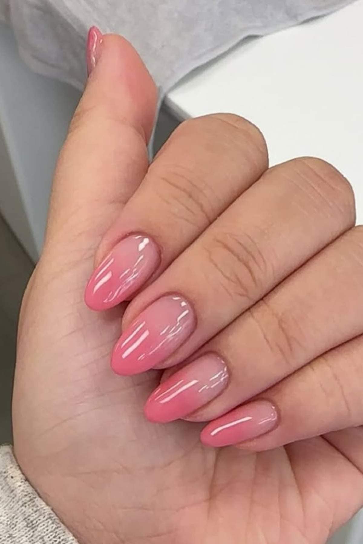 A hand with almond-shaped nails featuring a glossy pink ombre manicure, fading from light pink near the cuticle to a deeper pink at the tips.