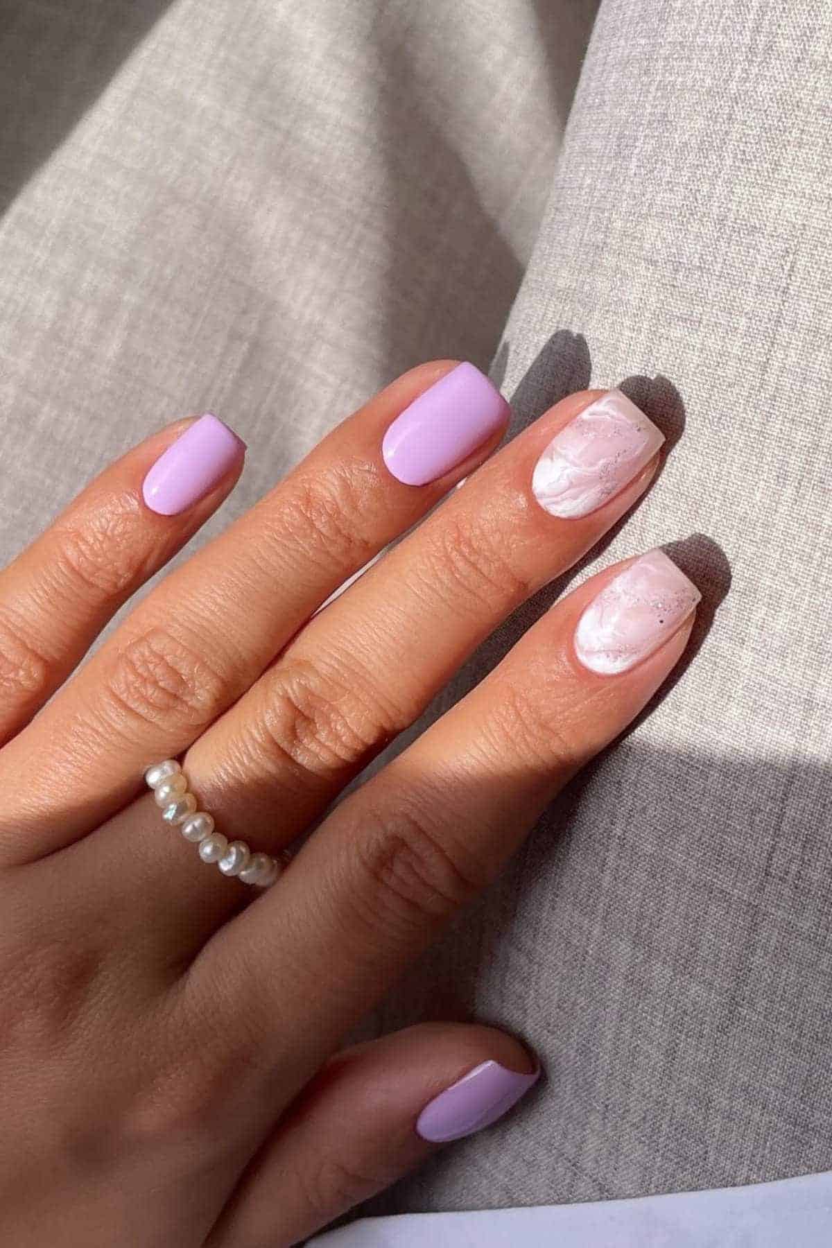 A hand with light purple nail polish on most nails and a marble design on the ring fingernail, wearing a delicate pearl ring.