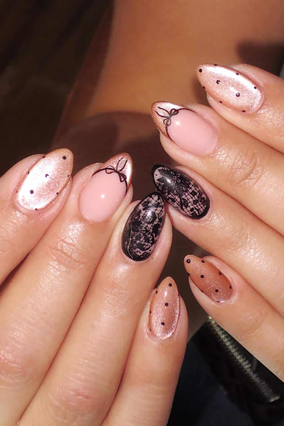 Close-up of hands with almond-shaped nails featuring nude, black lace, and glitter designs, with small bow and dot accents.